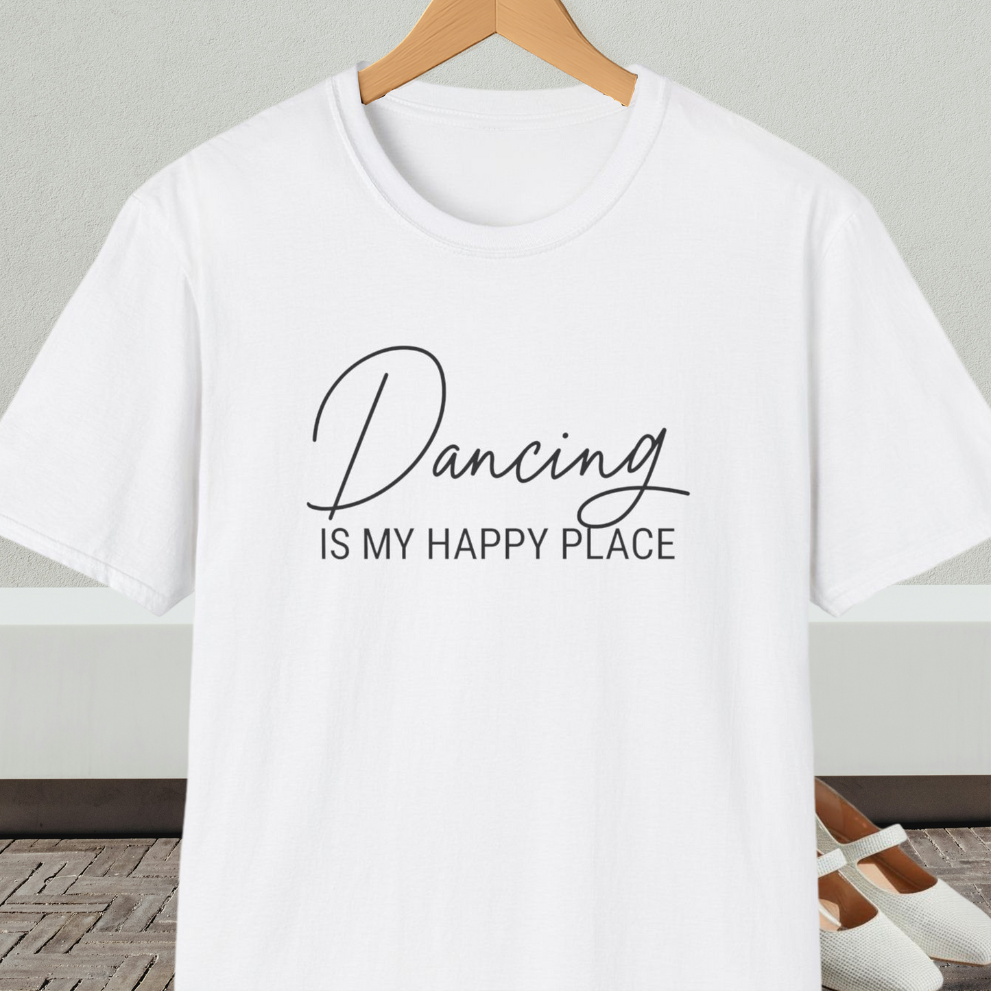 White t-shirt with 'Dancing is my happy place' text on a hanger against a neutral background.