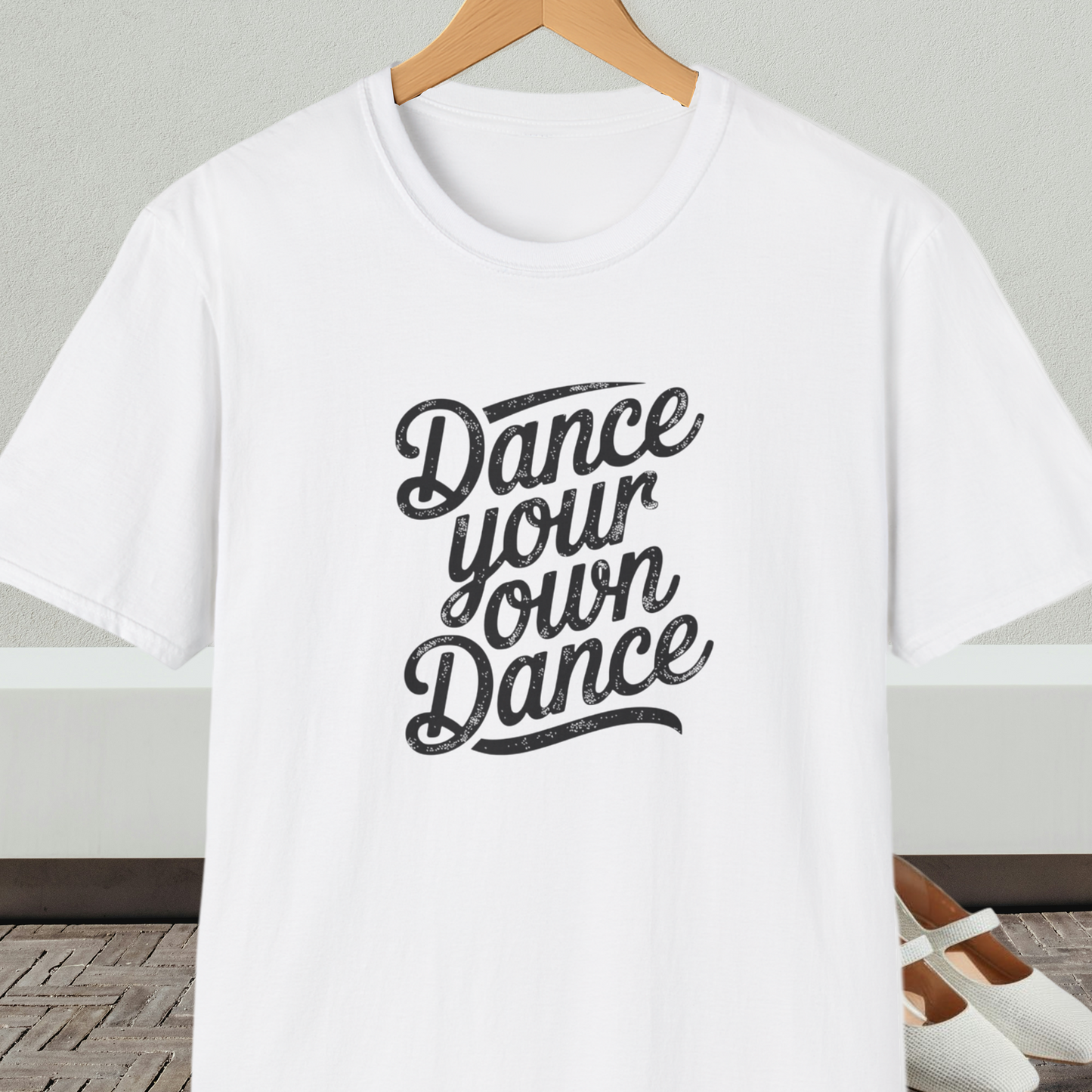 White t-shirt with 'Dance your own Dance' text, on a hanger against a neutral background.