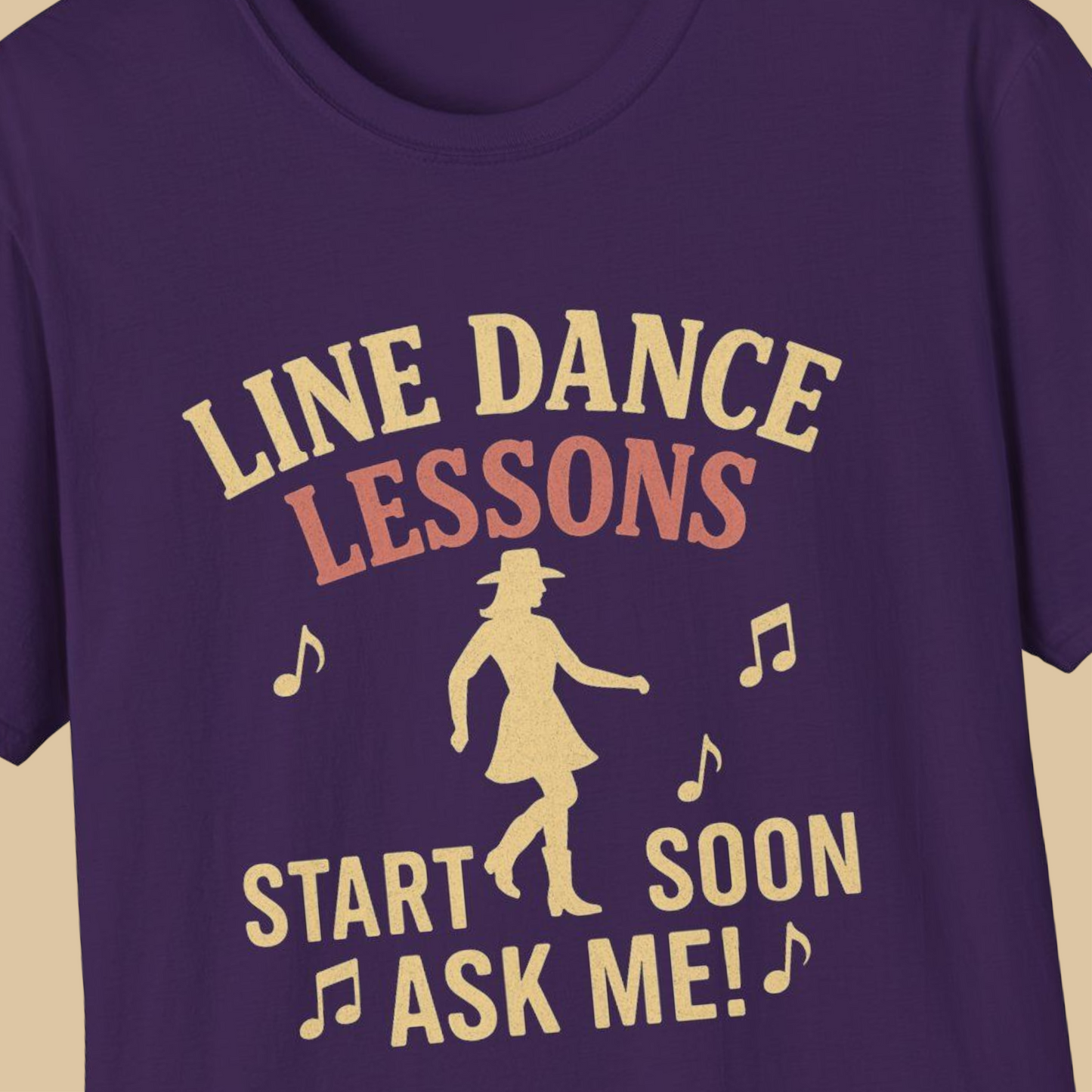A close-up of a purple t-shirt featuring a retro-style graphic print in a light tan color. The graphic reads 'LINE DANCE LESSONS START SOON ASK ME!' accompanied by musical notes and a silhouette of a woman in a cowboy hat and dress.