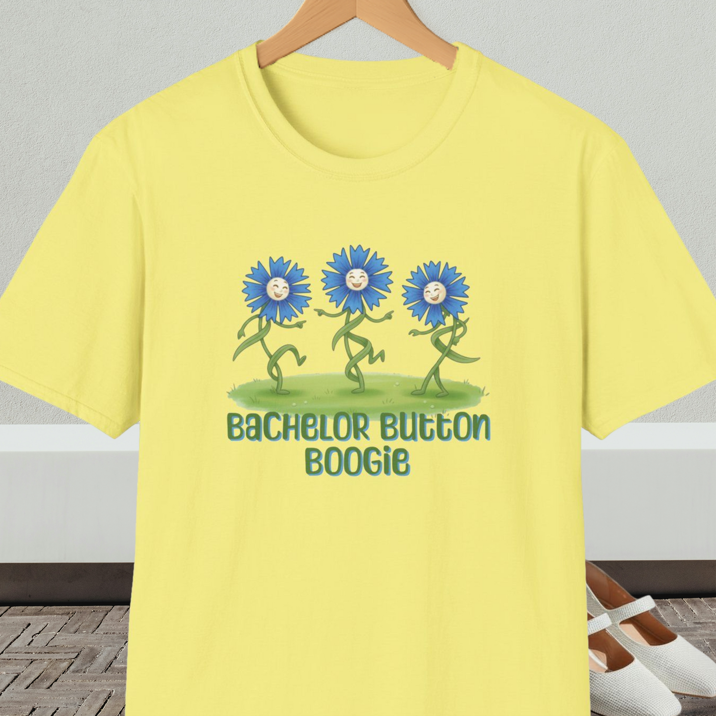 Yellow t-shirt with 'Bachelor Button Boogie' design featuring dancing blue flowers, on a hanger against a neutral background.