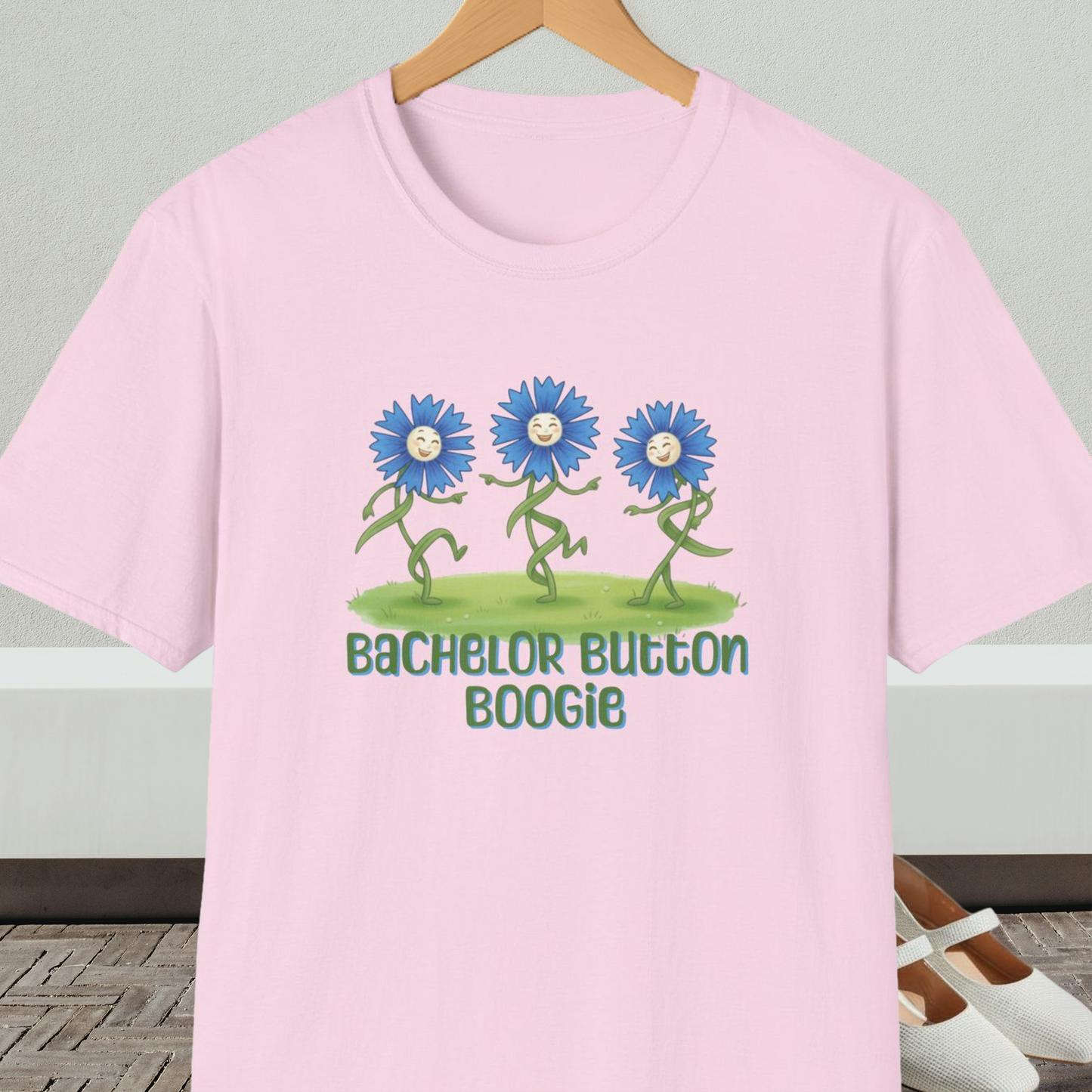 Light pink t-shirt with 'Bachelor Button Boogie' design featuring dancing blue flowers, on a hanger against a neutral background.