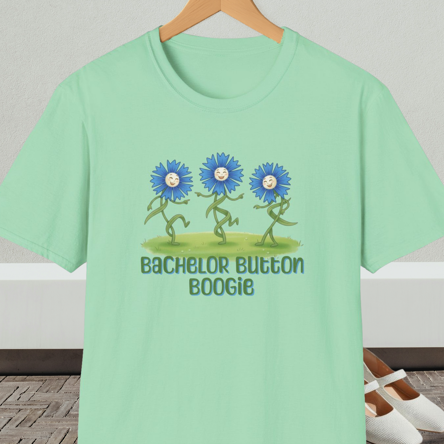 Light green t-shirt with 'Bachelor Button Boogie' design featuring dancing blue flowers, on a hanger against a neutral background.