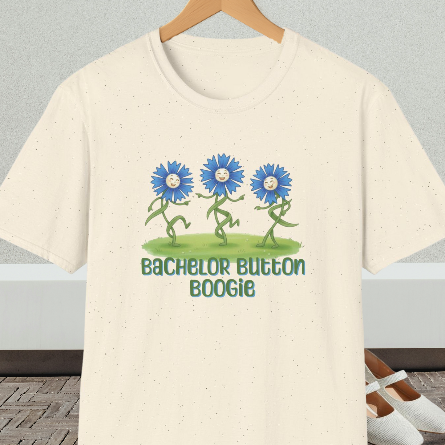 Natural beige t-shirt with 'Bachelor Button Boogie' design featuring dancing blue flowers, on a hanger against a neutral background.
