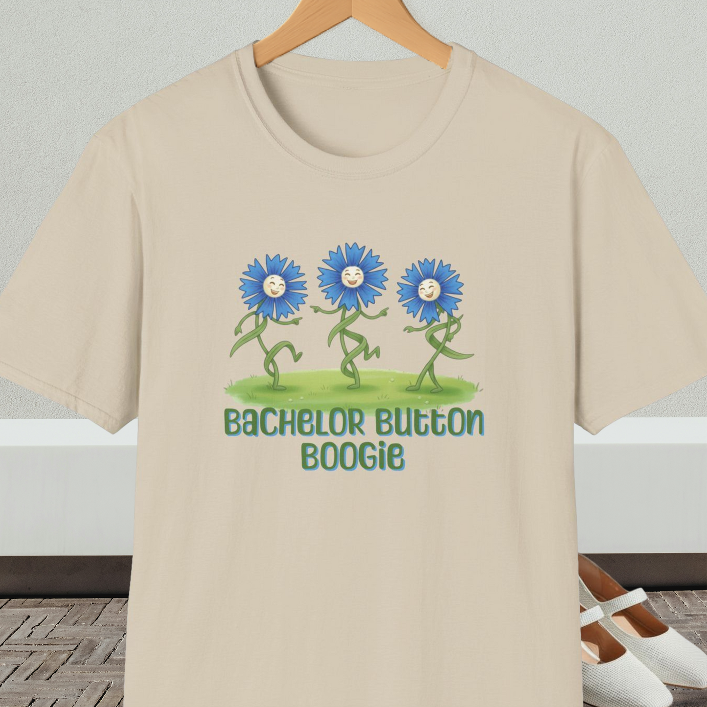 Beige t-shirt with 'Bachelor Button Boogie' design featuring dancing blue flowers, on a hanger against a neutral background.
