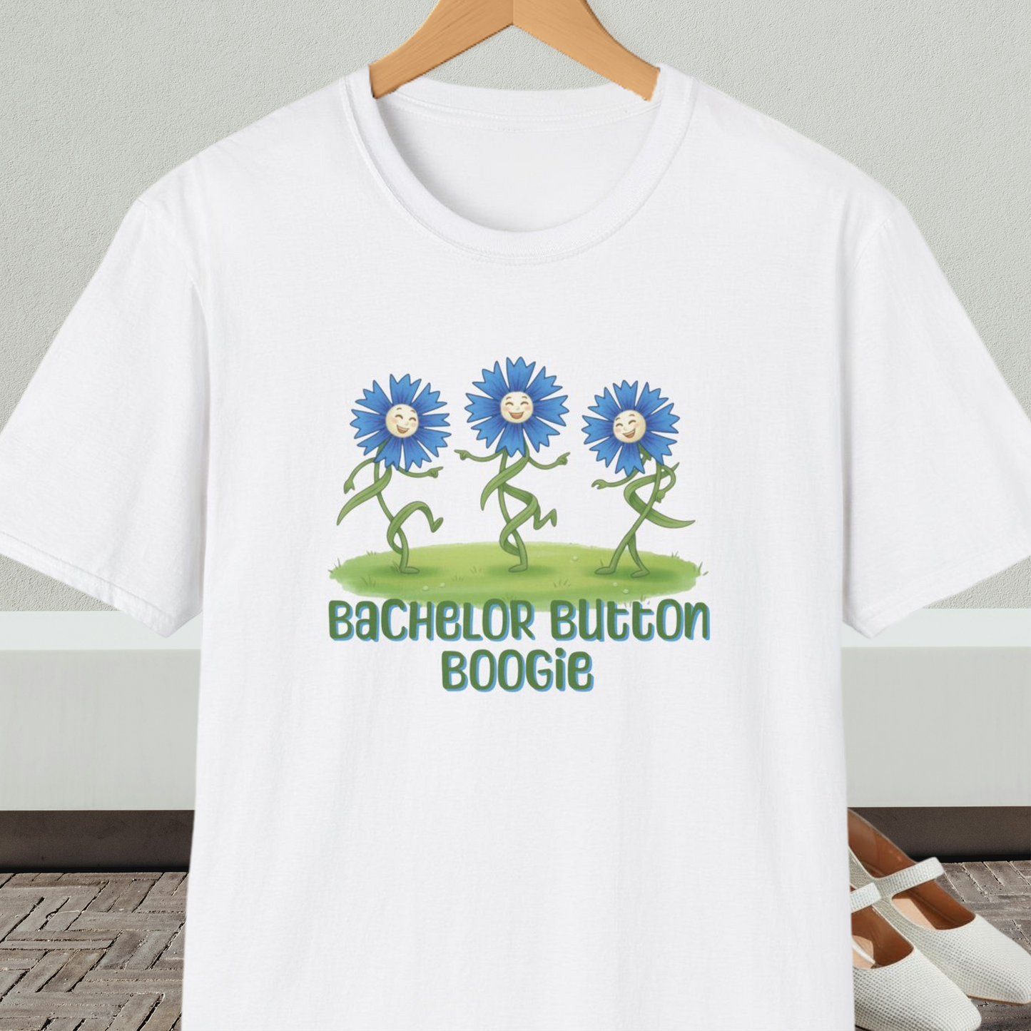 White t-shirt with 'Bachelor Button Boogie' design featuring dancing blue flowers, on a hanger against a neutral background.