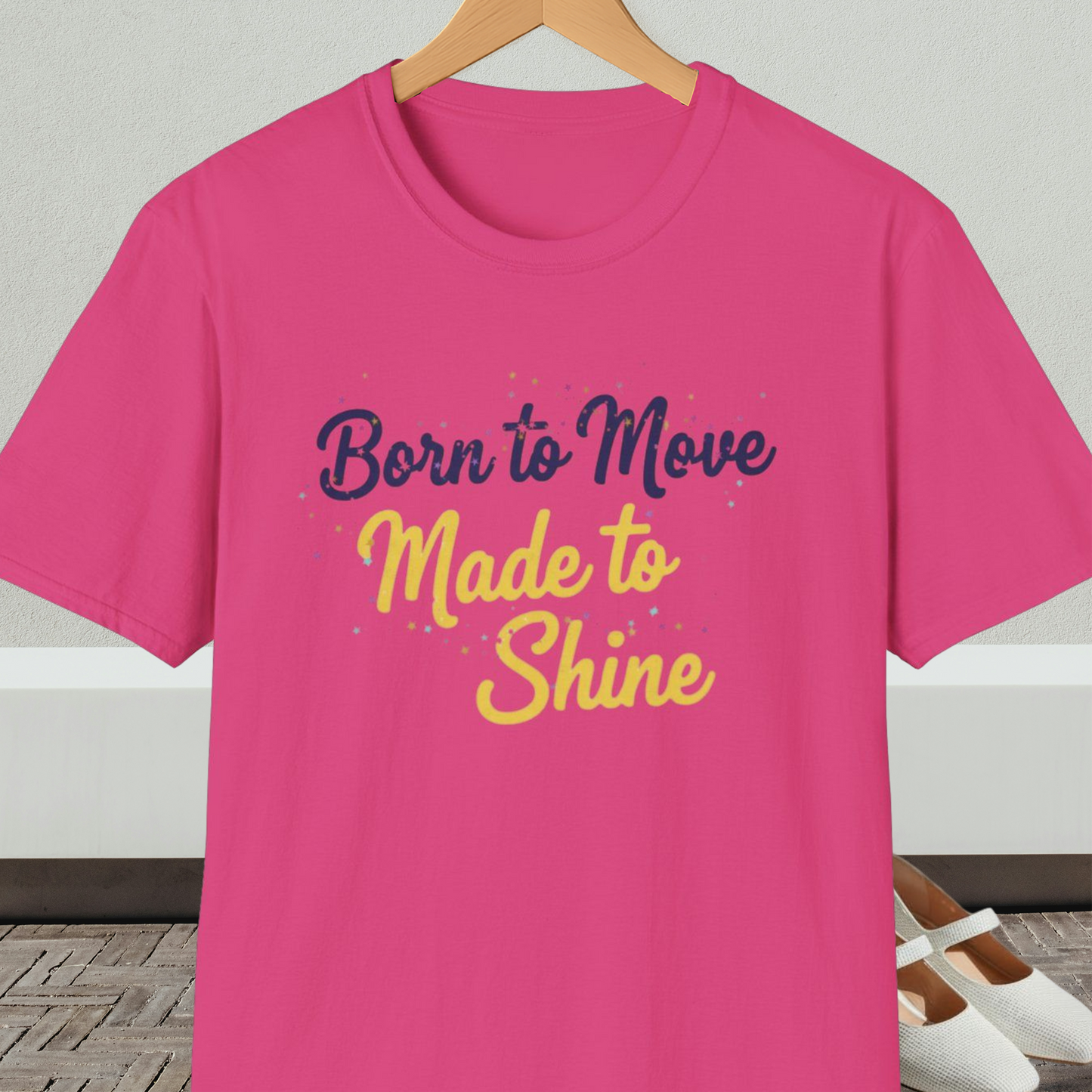 Bright pink t-shirt with 'Born to Move, Made to Shine' text, on a hanger against a white wall.