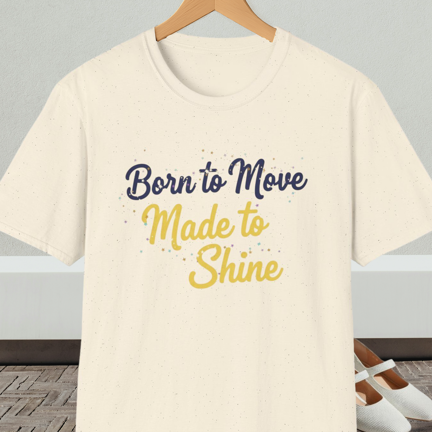 Natural beige t-shirt with 'Born to Move, Made to Shine' text, on a hanger against a white wall.