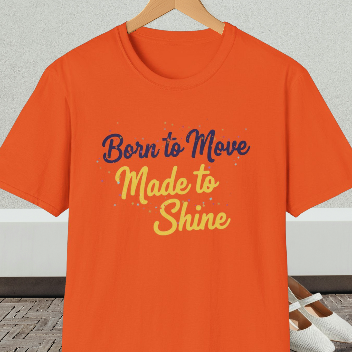 Orange t-shirt with 'Born to Move, Made to Shine' text, on a hanger against a white wall.