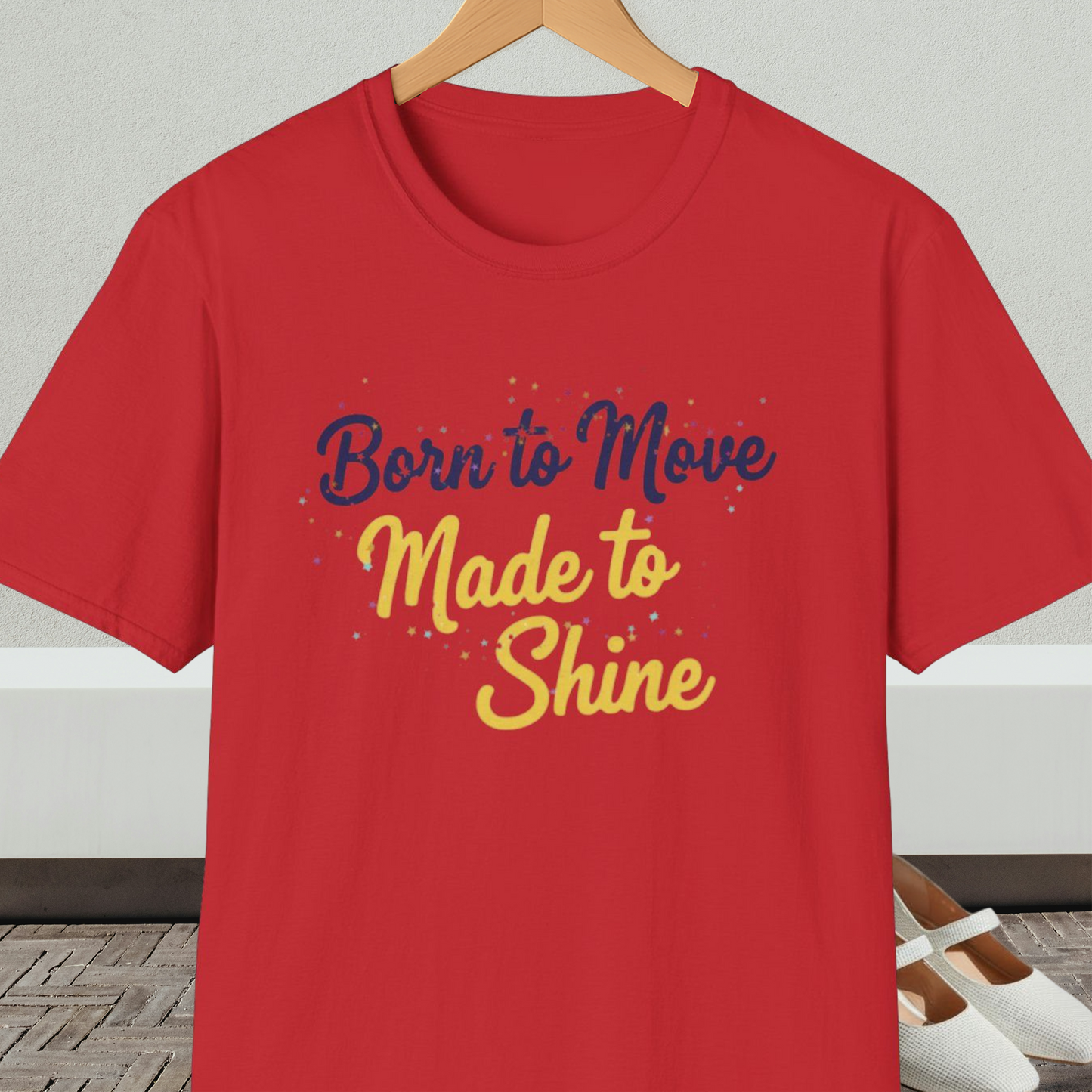 Red t-shirt with 'Born to Move, Made to Shine' text, on a hanger against a white wall.