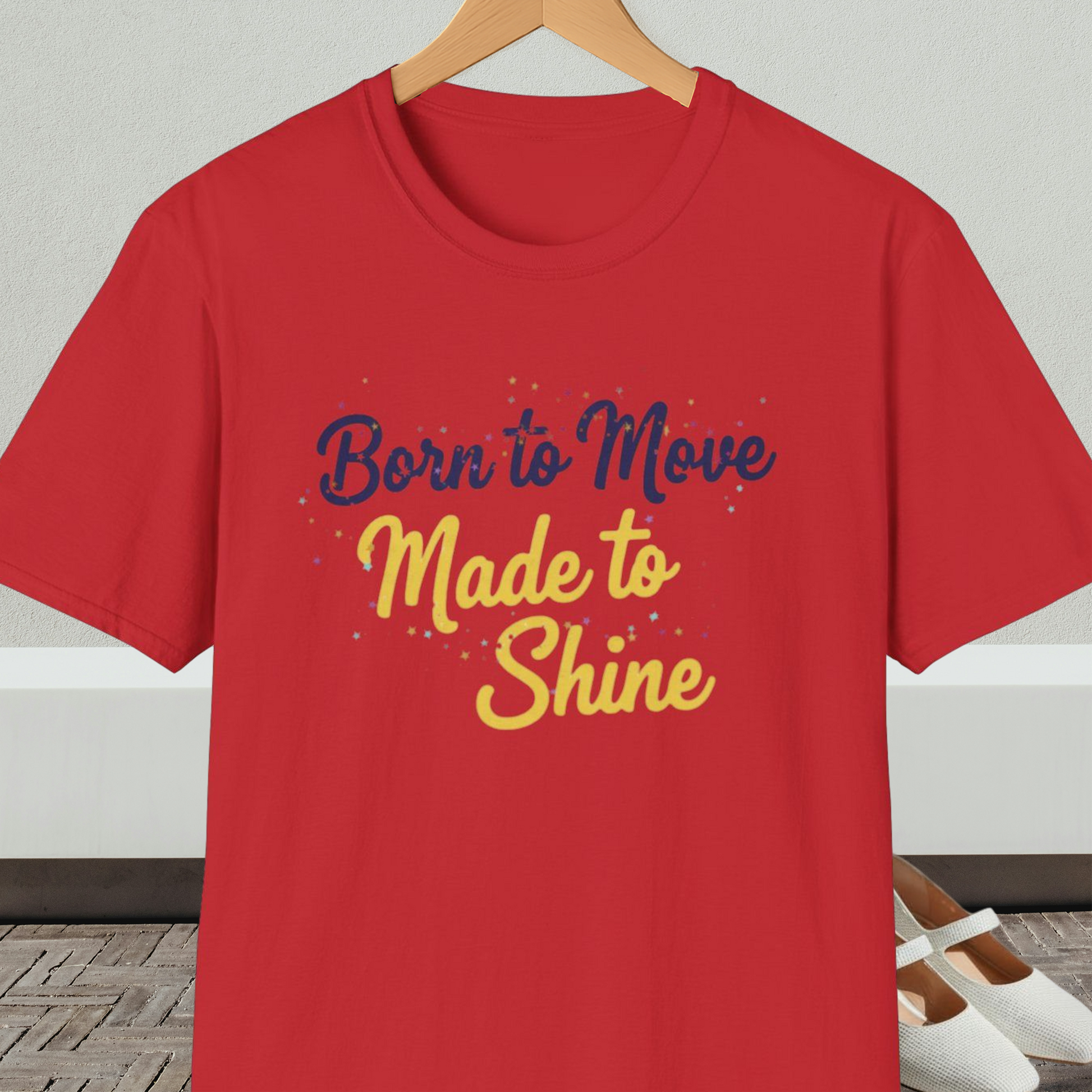 Red t-shirt with 'Born to Move, Made to Shine' text, on a hanger against a white wall.