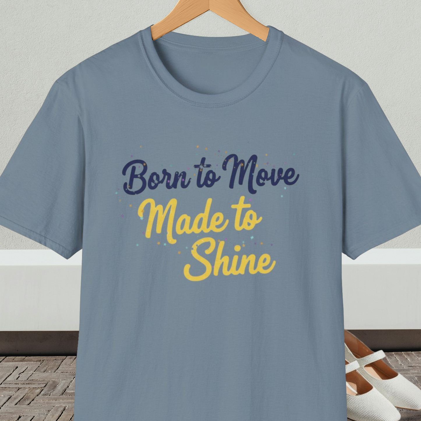 Stone blue t-shirt with 'Born to Move, Made to Shine' text, on a hanger against a white wall.