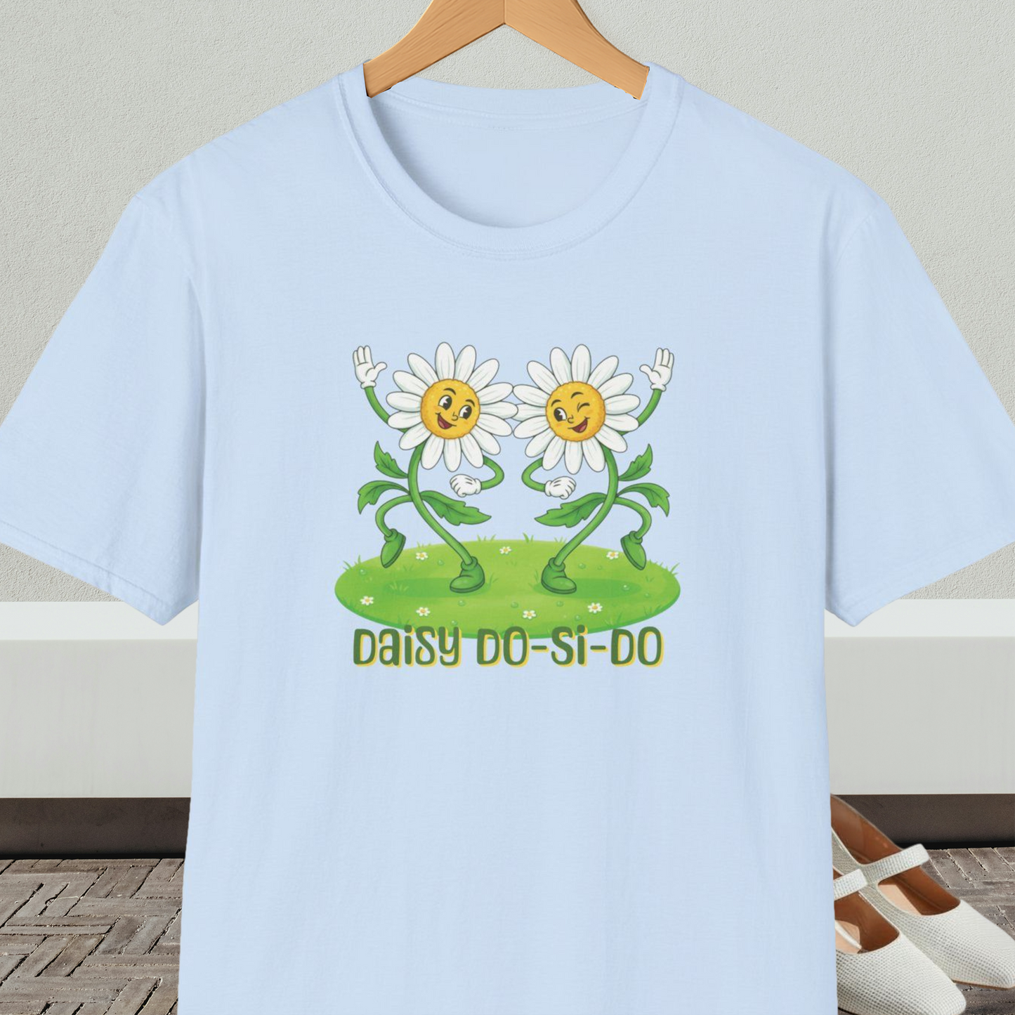Light blue t-shirt with cartoon daisies and text 'Daisy Do-Si-Do' on a wooden hanger against a white wall.
