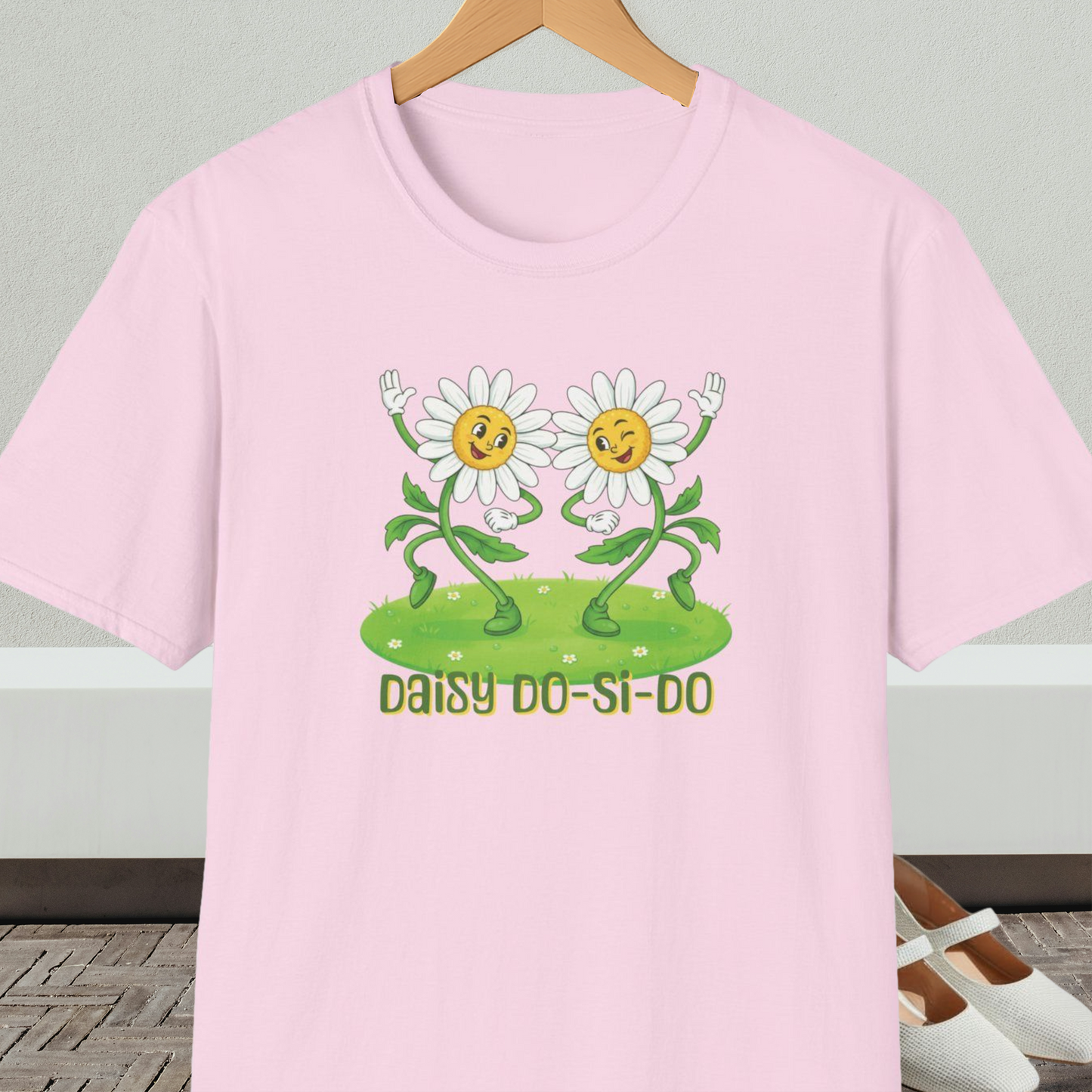 Light pink t-shirt with cartoon daisies and text 'Daisy Do-Si-Do' on a wooden hanger against a white wall.