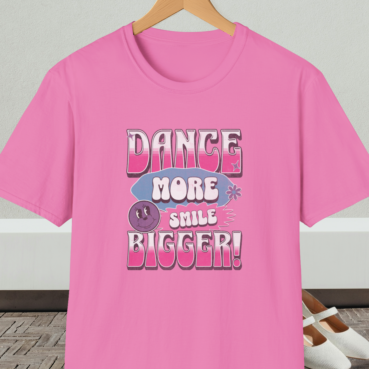 Pink t-shirt with 'Dance More Smile Bigger!' retro text, on a hanger against a neutral background.
