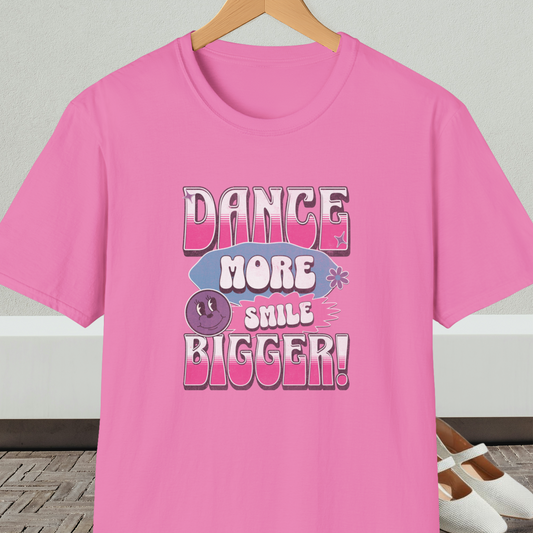 Pink t-shirt with 'Dance More Smile Bigger!' retro text, on a hanger against a neutral background.