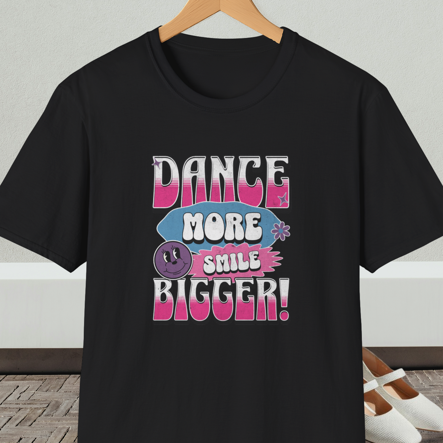 Black t-shirt with 'Dance More Smile Bigger!' retro text, on a hanger against a neutral background.