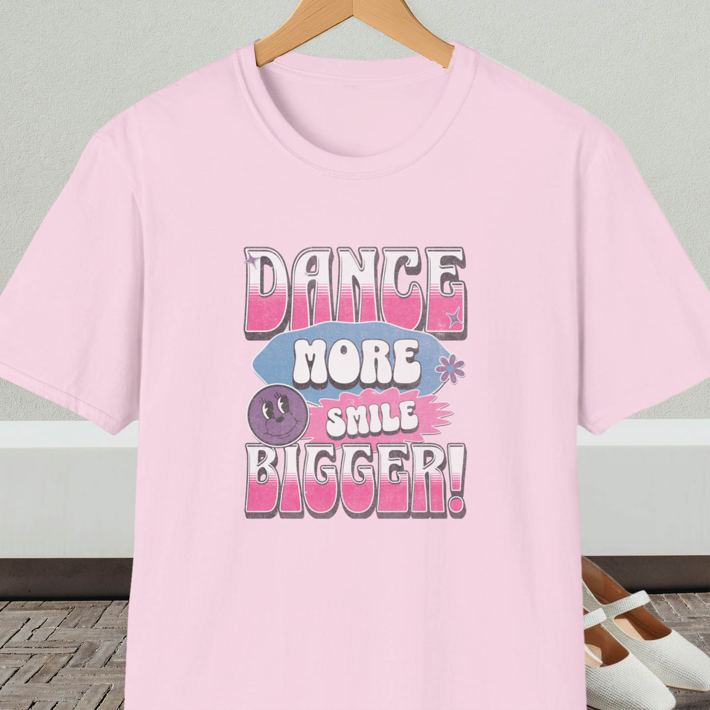 Light pink t-shirt with 'Dance More Smile Bigger!' retro text, on a hanger against a neutral background.