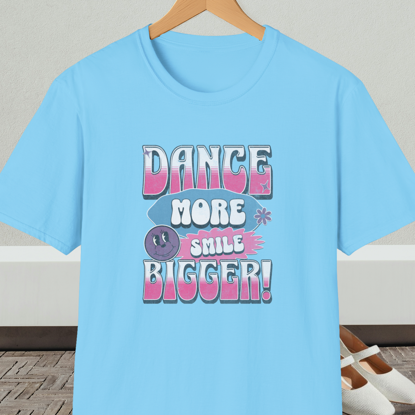 Light blue t-shirt with 'Dance More Smile Bigger!' retro text, on a hanger against a neutral background.
