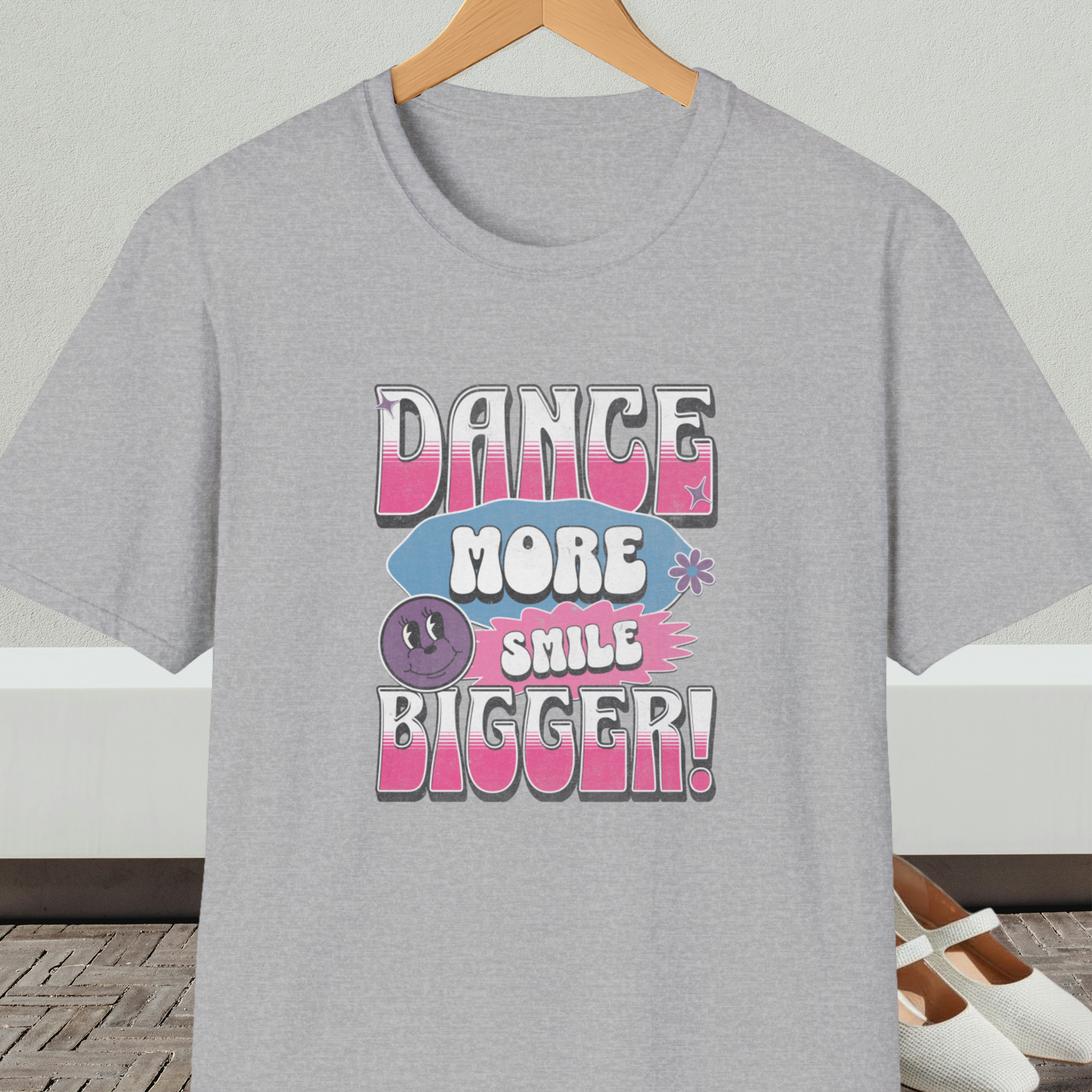 Gray t-shirt with 'Dance More Smile Bigger!' retro text, on a hanger against a neutral background.