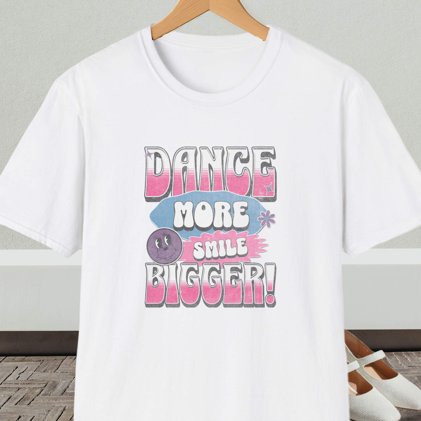 White t-shirt with 'Dance More Smile Bigger!' retro text, on a hanger against a neutral background.