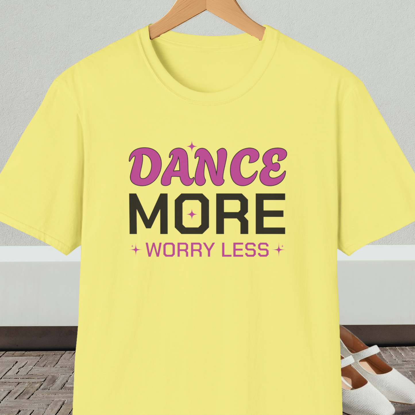 Yellow t-shirt with 'Dance More Worry Less' text, on a hanger against a neutral background