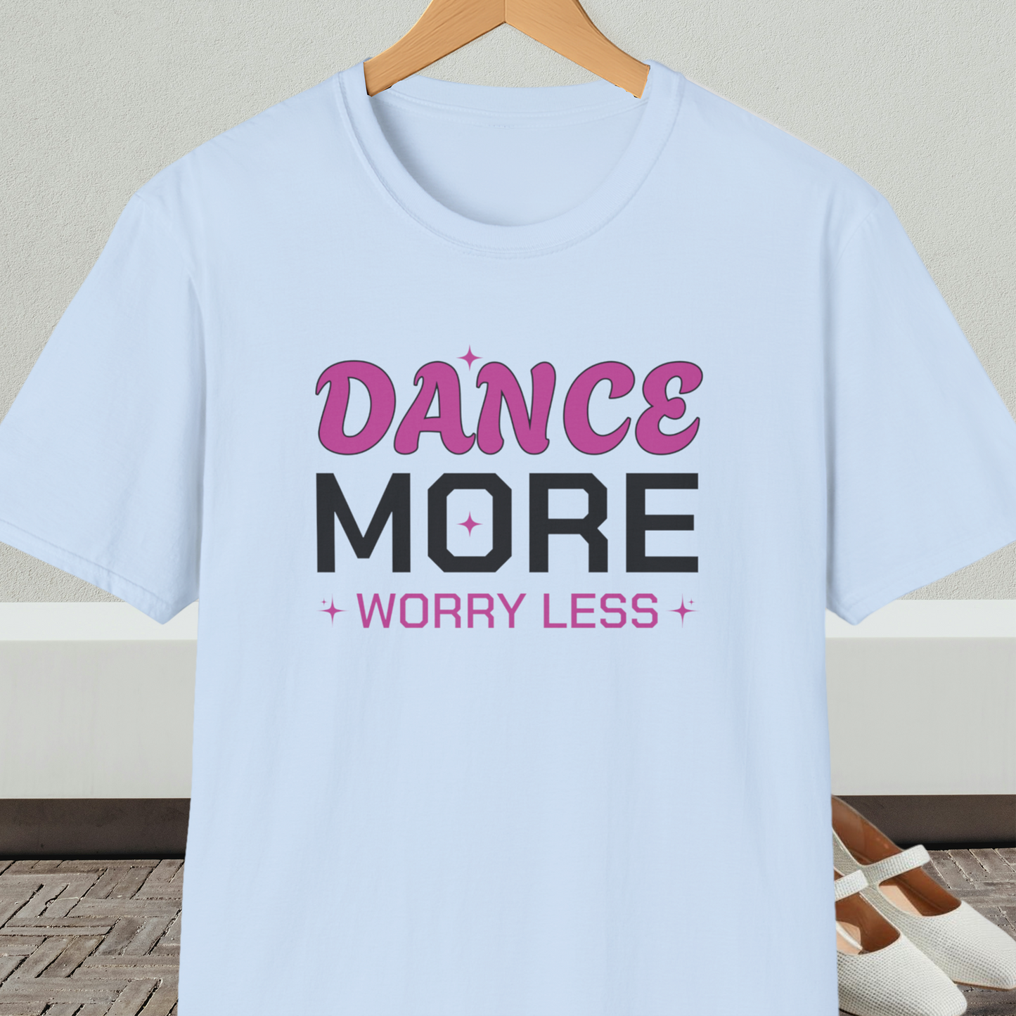 Light blue t-shirt with 'Dance More Worry Less' text, on a hanger against a neutral background