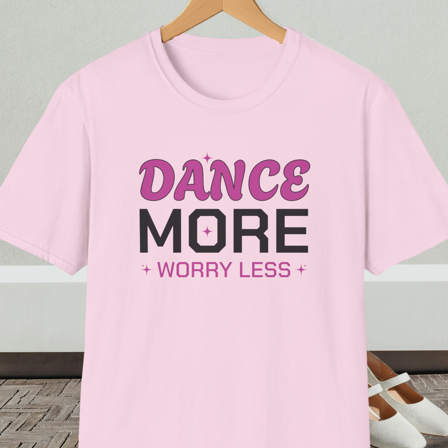 Light pink t-shirt with 'Dance More Worry Less' text, on a hanger against a neutral background