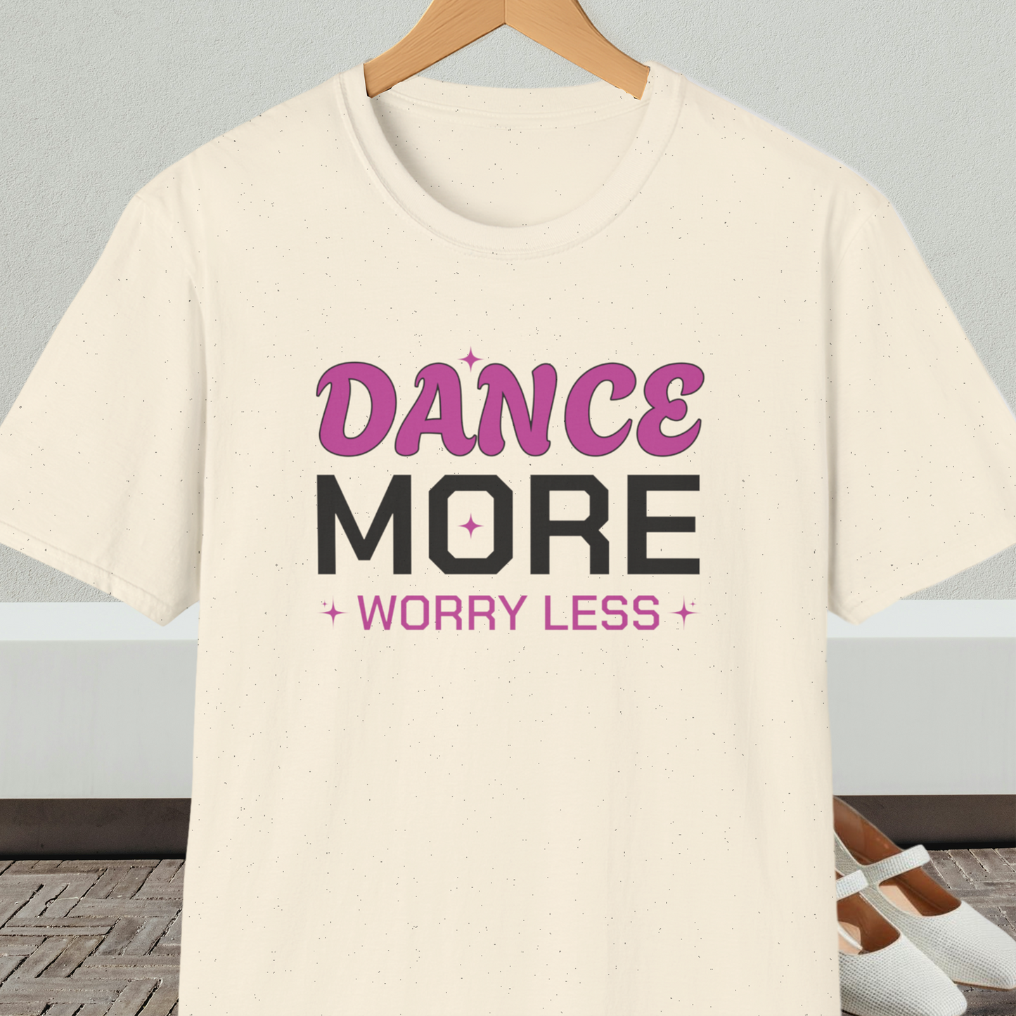 Natural beige t-shirt with 'Dance More Worry Less' text, on a hanger against a neutral background