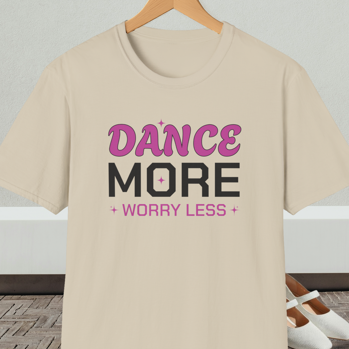 Beige t-shirt with 'Dance More Worry Less' text, on a hanger against a neutral background