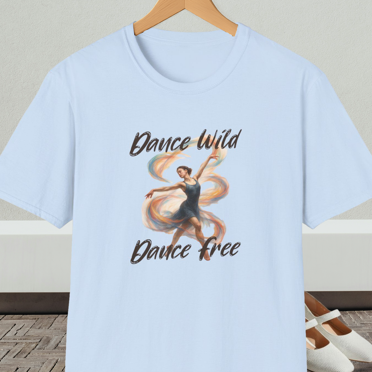 Light blue t-shirt with a graphic of a dancer and text 'Dance Wild, Dance Free', on a hanger against a neutral background.