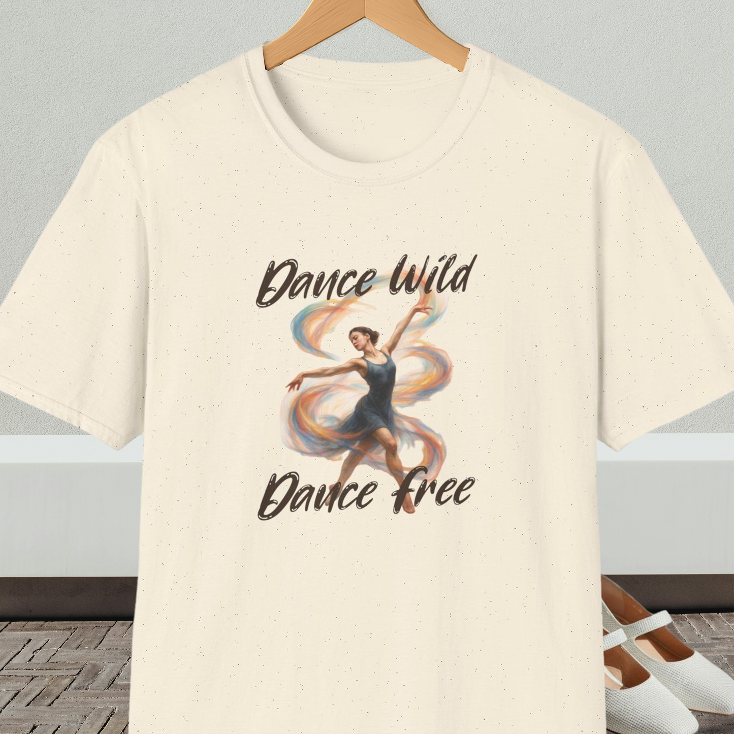 Natural beige t-shirt with a graphic of a dancer and text 'Dance Wild, Dance Free', on a hanger against a neutral background.