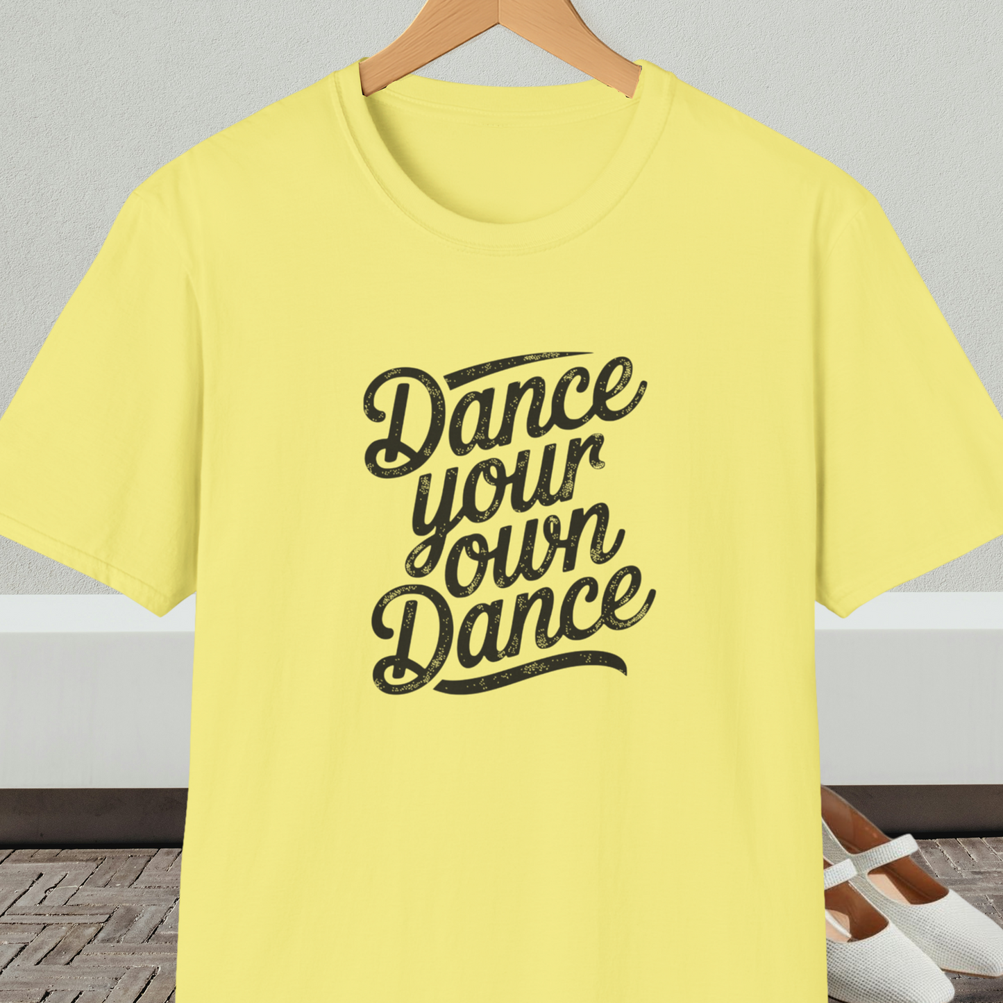Yellow t-shirt with 'Dance your own Dance' text, on a hanger against a neutral background.