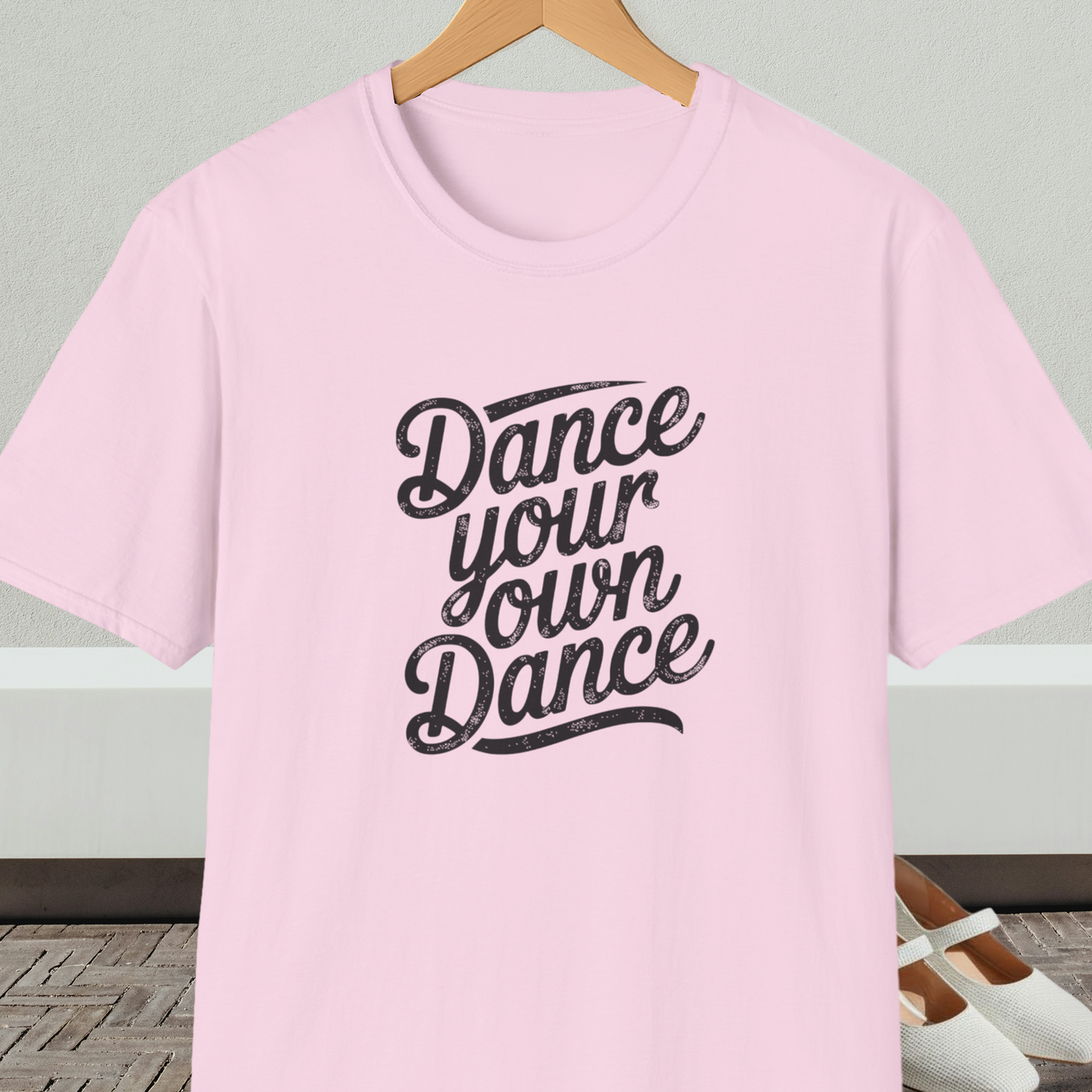 Light pink t-shirt with 'Dance your own Dance' text, on a hanger against a neutral background.