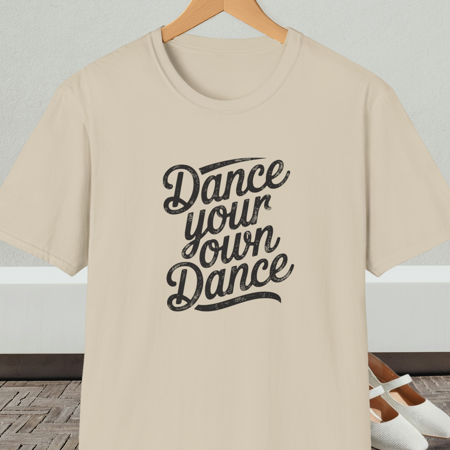 Beige t-shirt with 'Dance your own Dance' text, on a hanger against a neutral background.