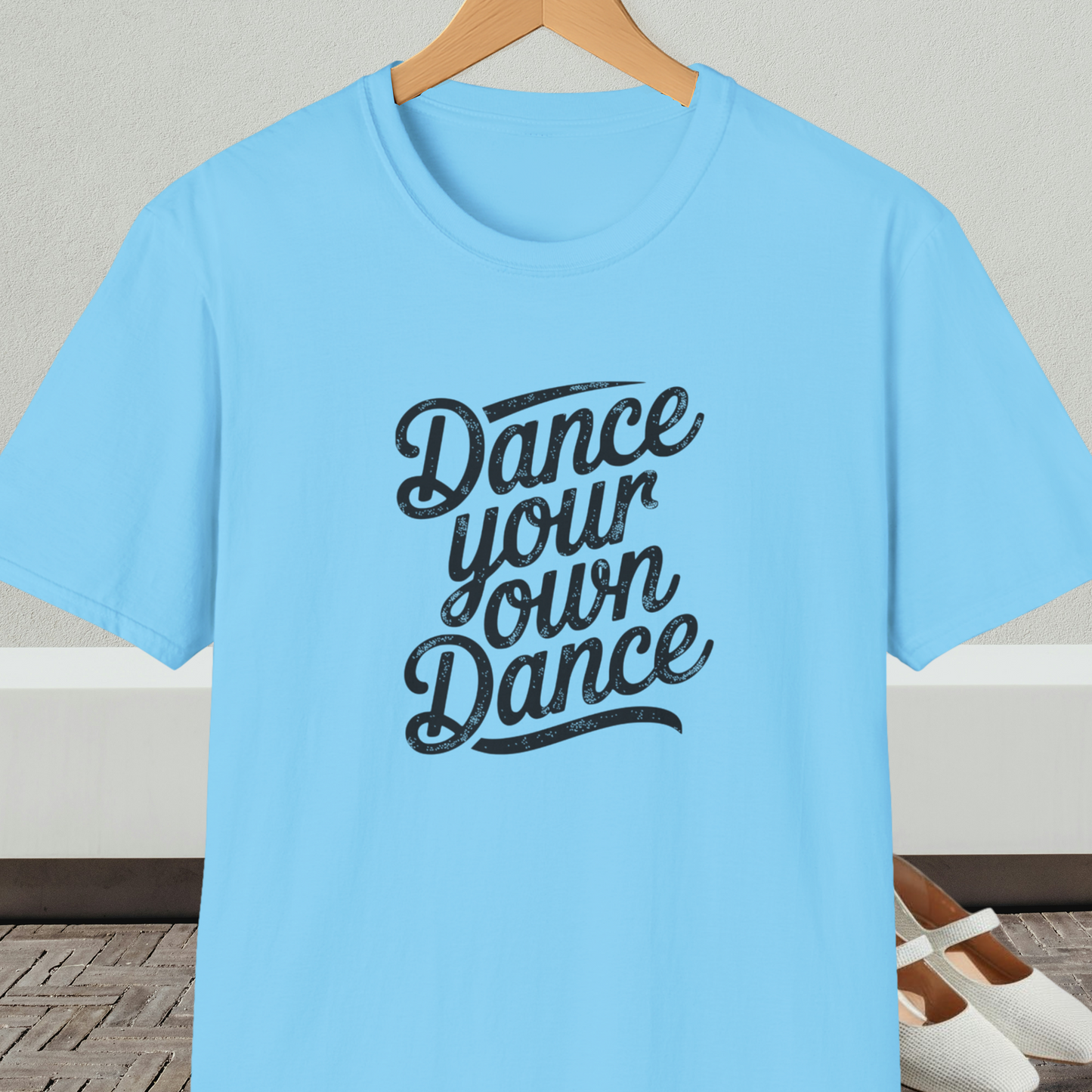 Light blue t-shirt with 'Dance your own Dance' text, on a hanger against a neutral background.