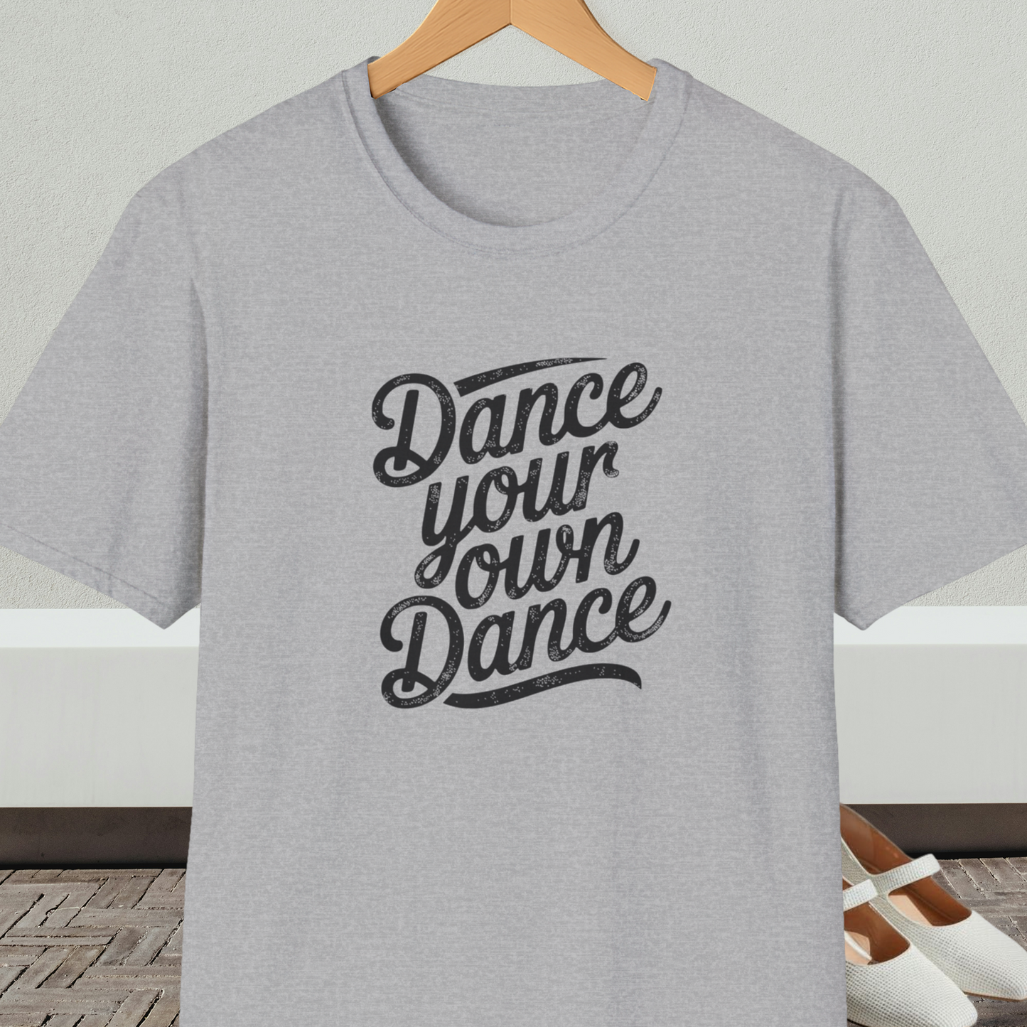 Gray t-shirt with 'Dance your own Dance' text, on a hanger against a neutral background.