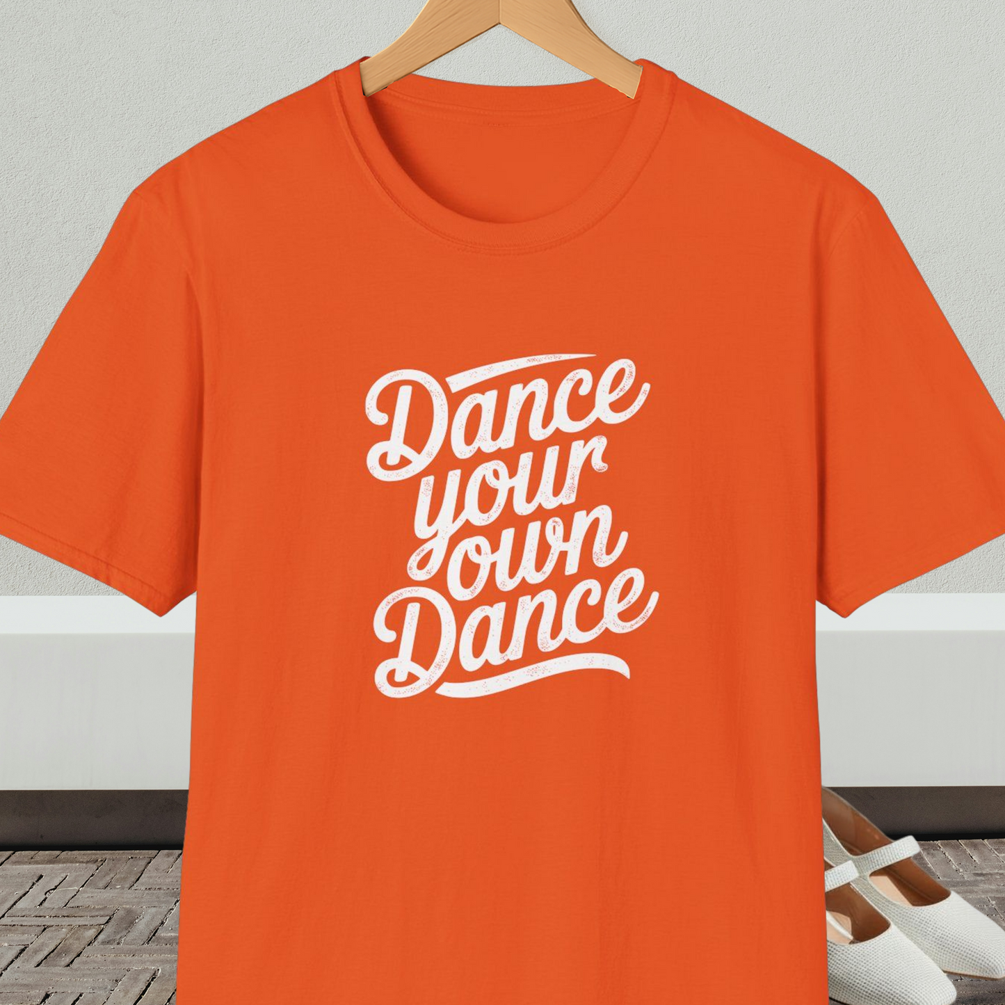 Orange t-shirt with 'Dance your own Dance' text, on a hanger against a neutral background
