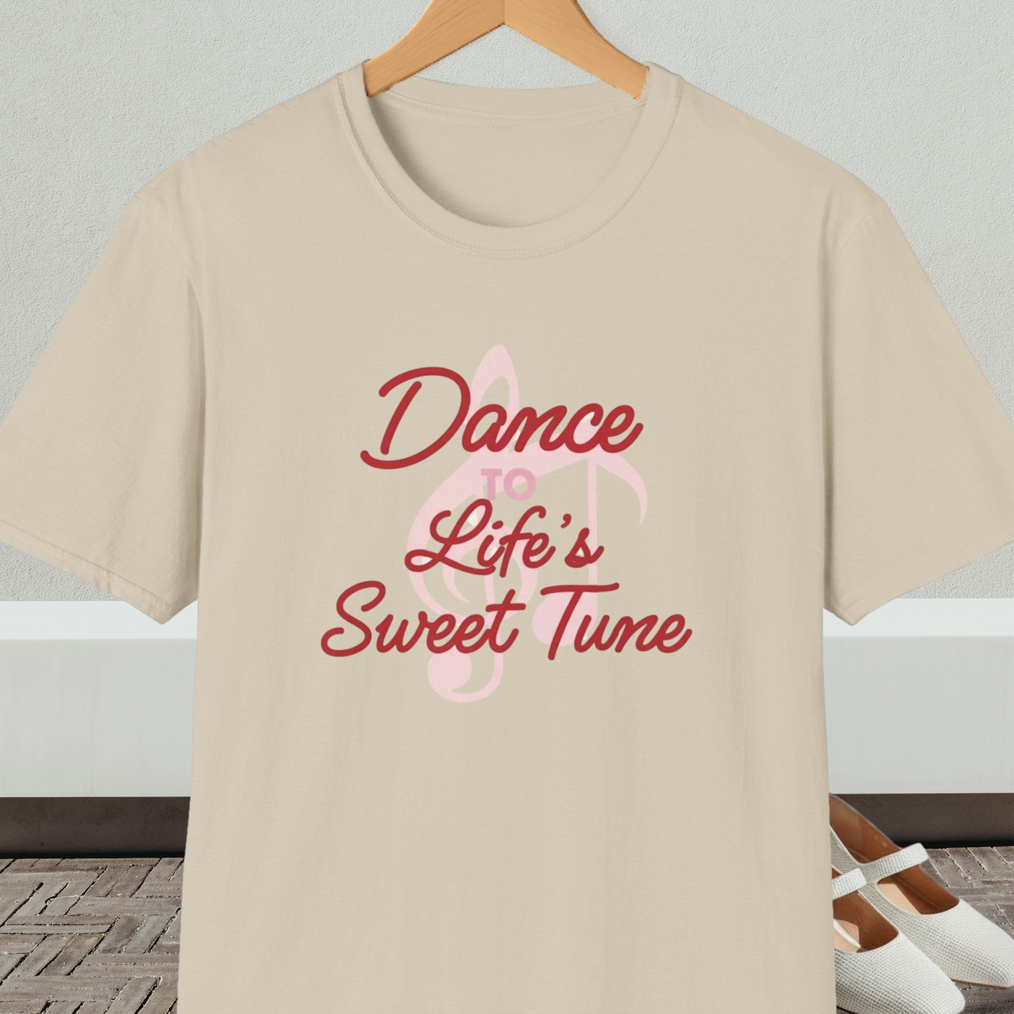 Beige t-shirt with 'Dance to Life's Sweet Tune' text and pink musical note design, on a hanger against a neutral background.