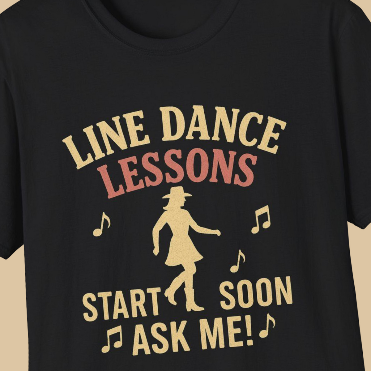 A close-up of a black t-shirt featuring a retro-style graphic print in a light tan color. The graphic reads 'LINE DANCE LESSONS START SOON ASK ME!' accompanied by musical notes and a silhouette of a woman in a cowboy hat and dress.