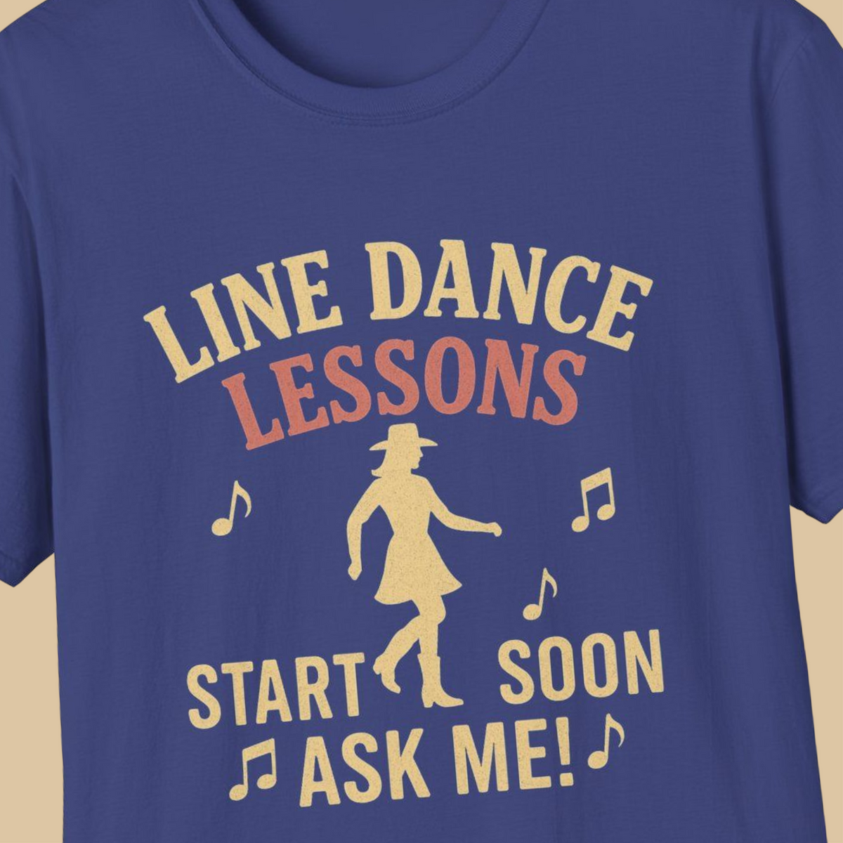 A close-up of a blue t-shirt featuring a retro-style graphic print in a light tan color. The graphic reads 'LINE DANCE LESSONS START SOON ASK ME!' accompanied by musical notes and a silhouette of a woman in a cowboy hat and dress.
