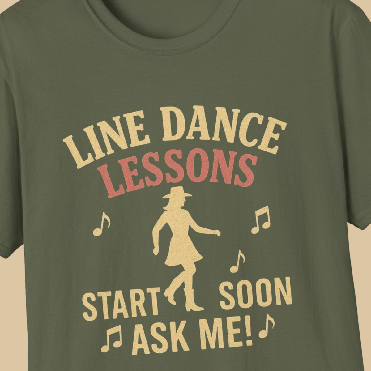 A close-up of a military green t-shirt featuring a retro-style graphic print in a light tan color. The graphic reads 'LINE DANCE LESSONS START SOON ASK ME!' accompanied by musical notes and a silhouette of a woman in a cowboy hat and dress.