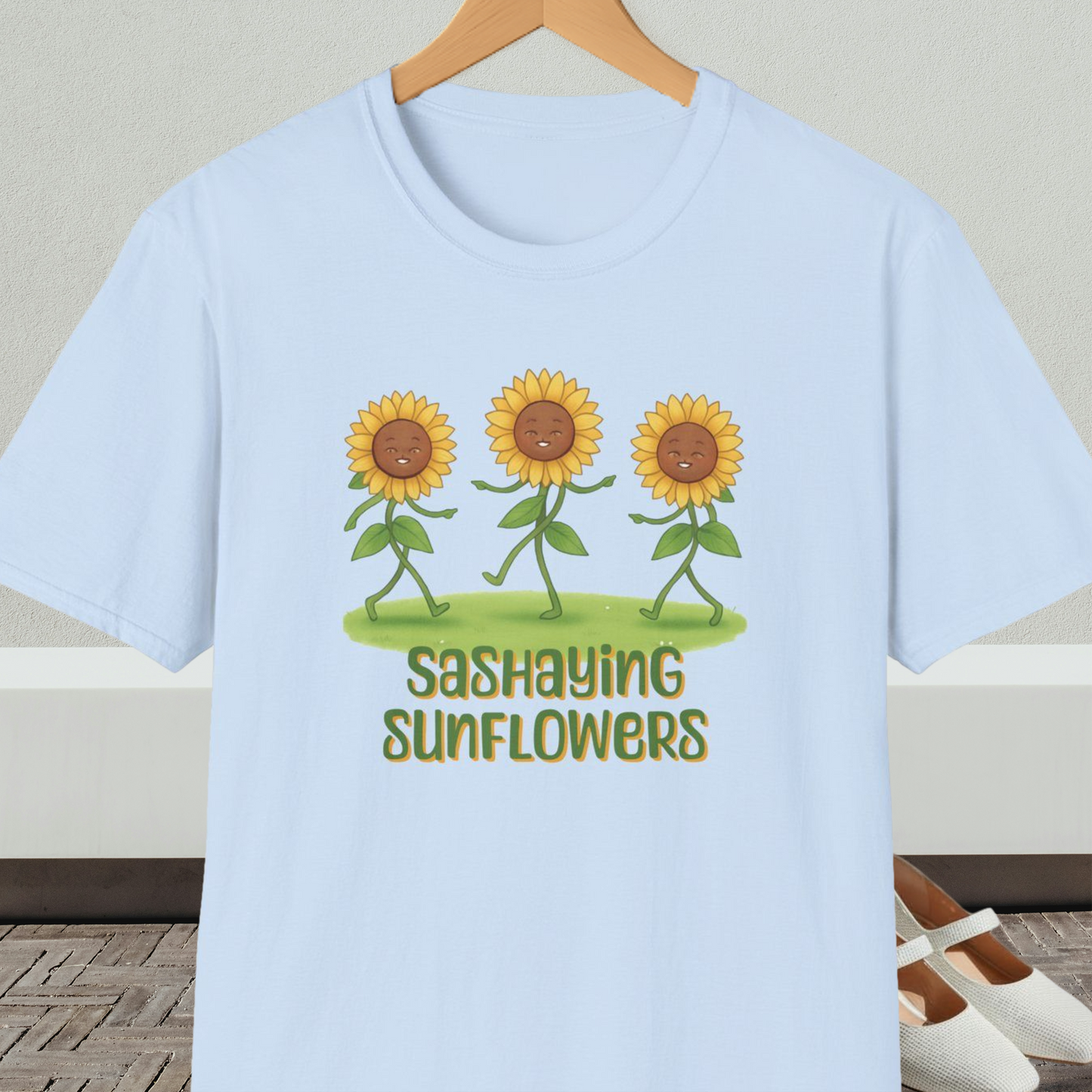 Light blue t-shirt with cartoon sunflowers and 'Sashaying Sunflowers' text, on a hanger against a neutral background.