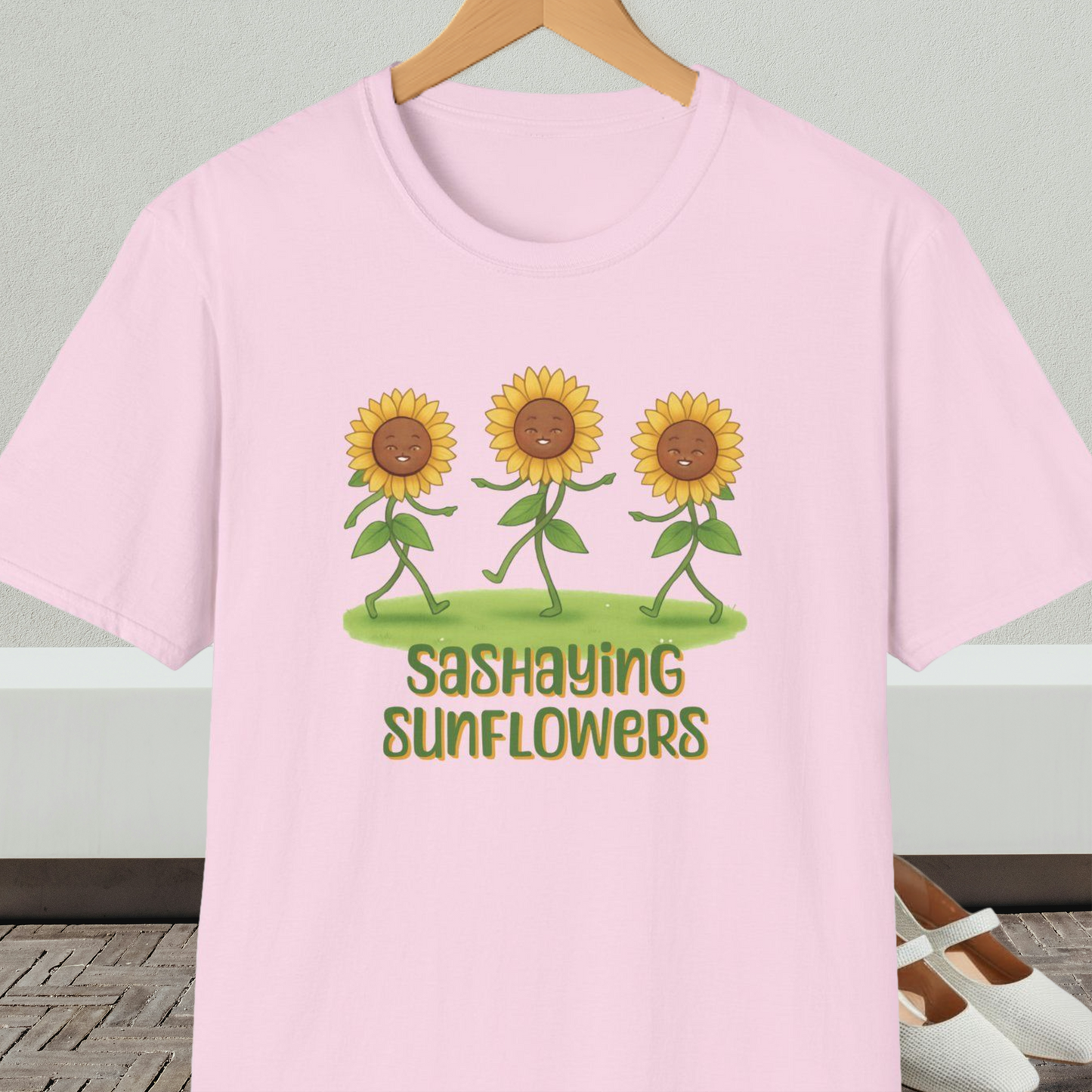 Light pink t-shirt with cartoon sunflowers and 'Sashaying Sunflowers' text, on a hanger against a neutral background.