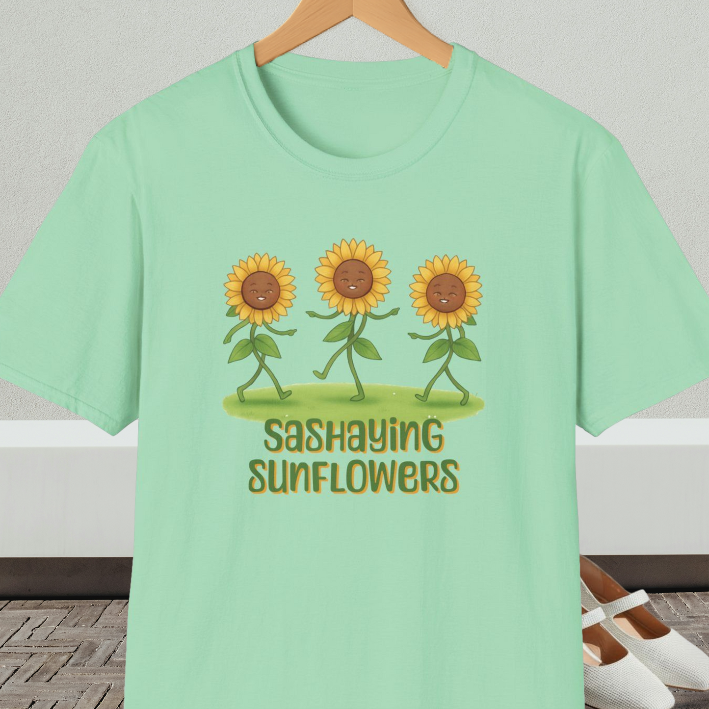 Mint green t-shirt with cartoon sunflowers and 'Sashaying Sunflowers' text, on a hanger against a neutral background.