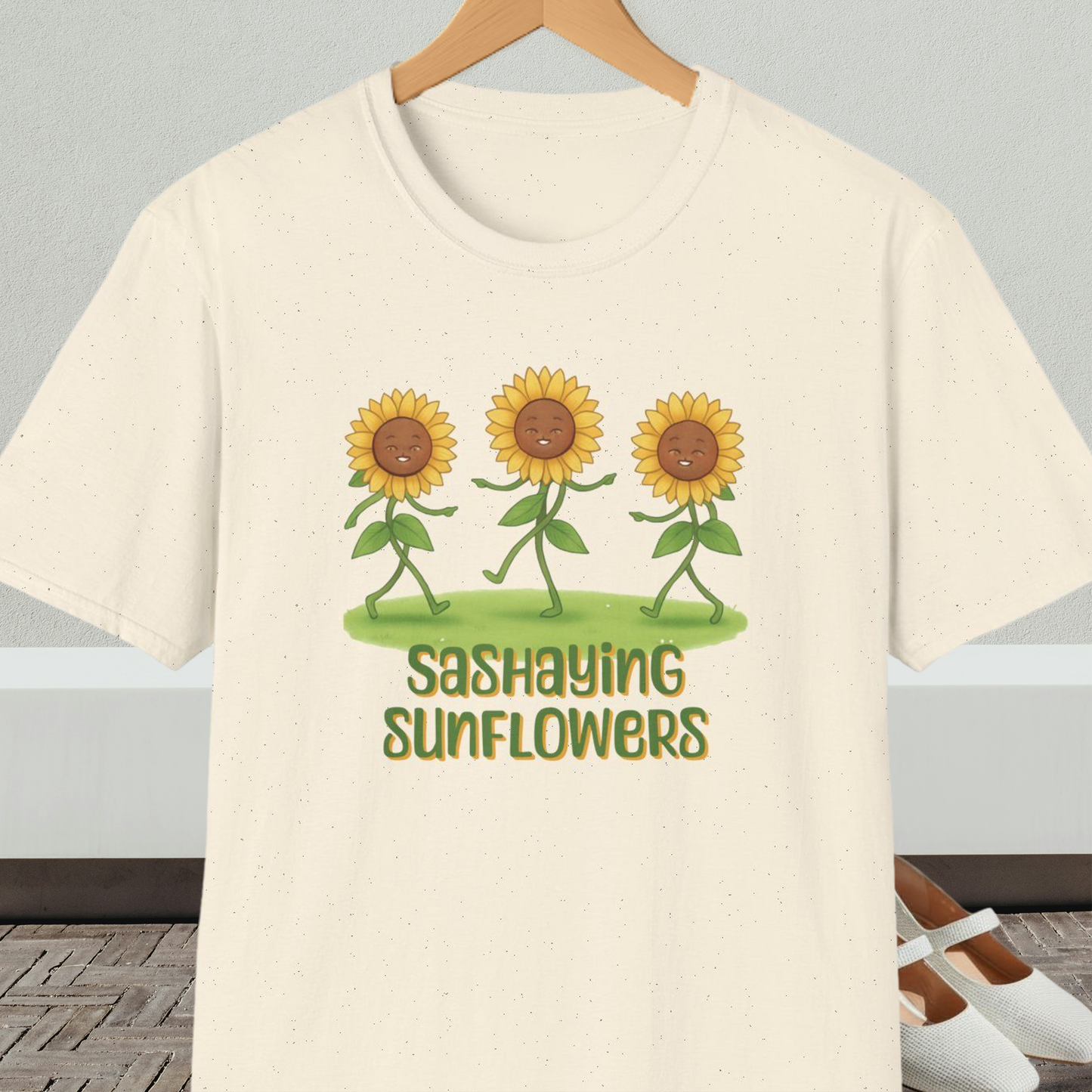 Natural beige t-shirt with cartoon sunflowers and 'Sashaying Sunflowers' text, on a hanger against a neutral background.