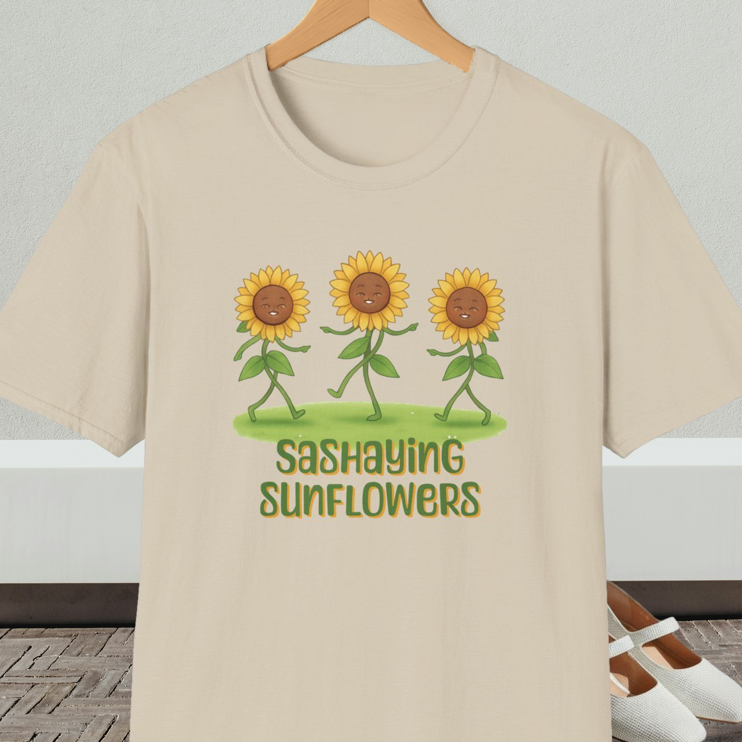 Beige t-shirt with cartoon sunflowers and 'Sashaying Sunflowers' text, on a hanger against a neutral background.