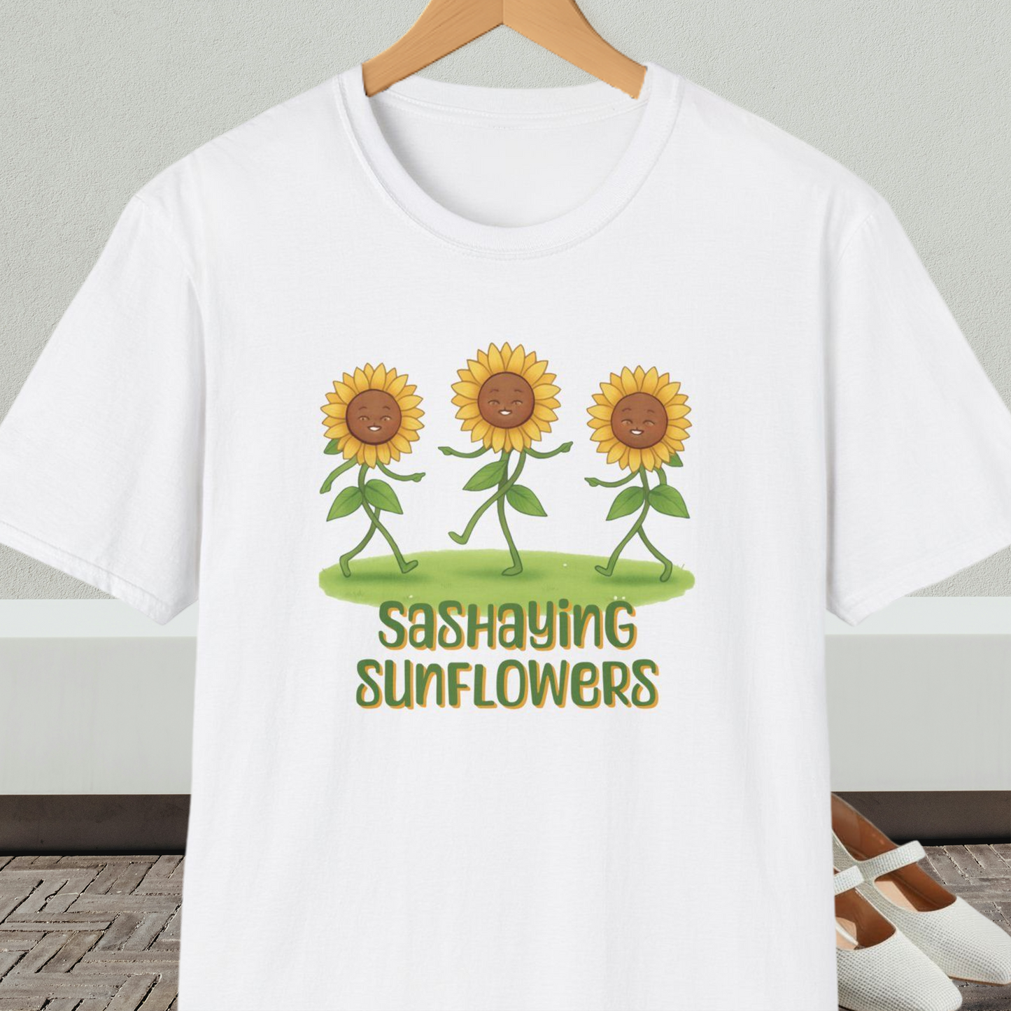 White t-shirt with cartoon sunflowers and 'Sashaying Sunflowers' text, on a hanger against a neutral background.