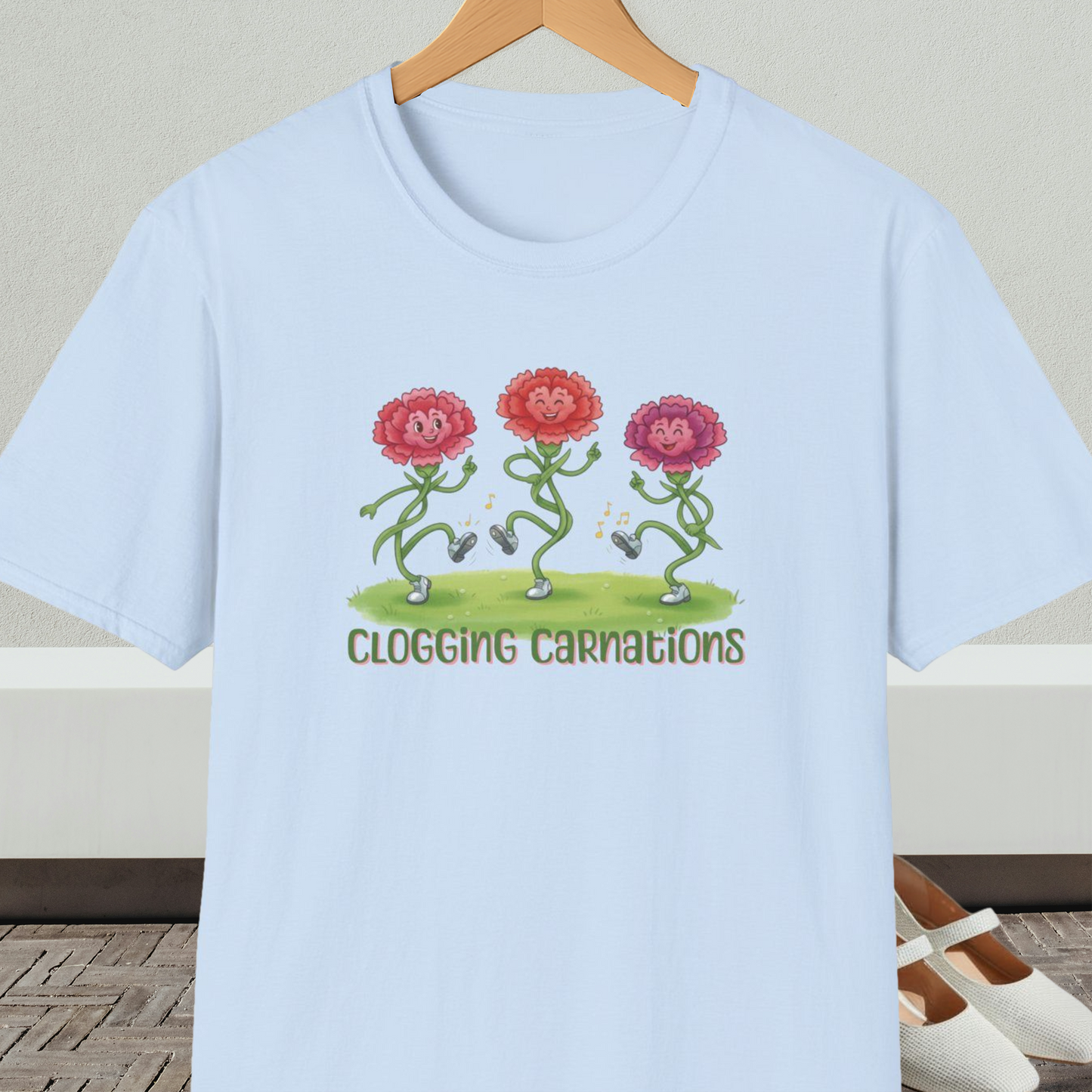 Light blue t-shirt with dancing flower design and text 'Clogging Carnations', on a hanger in front of a neutral background.