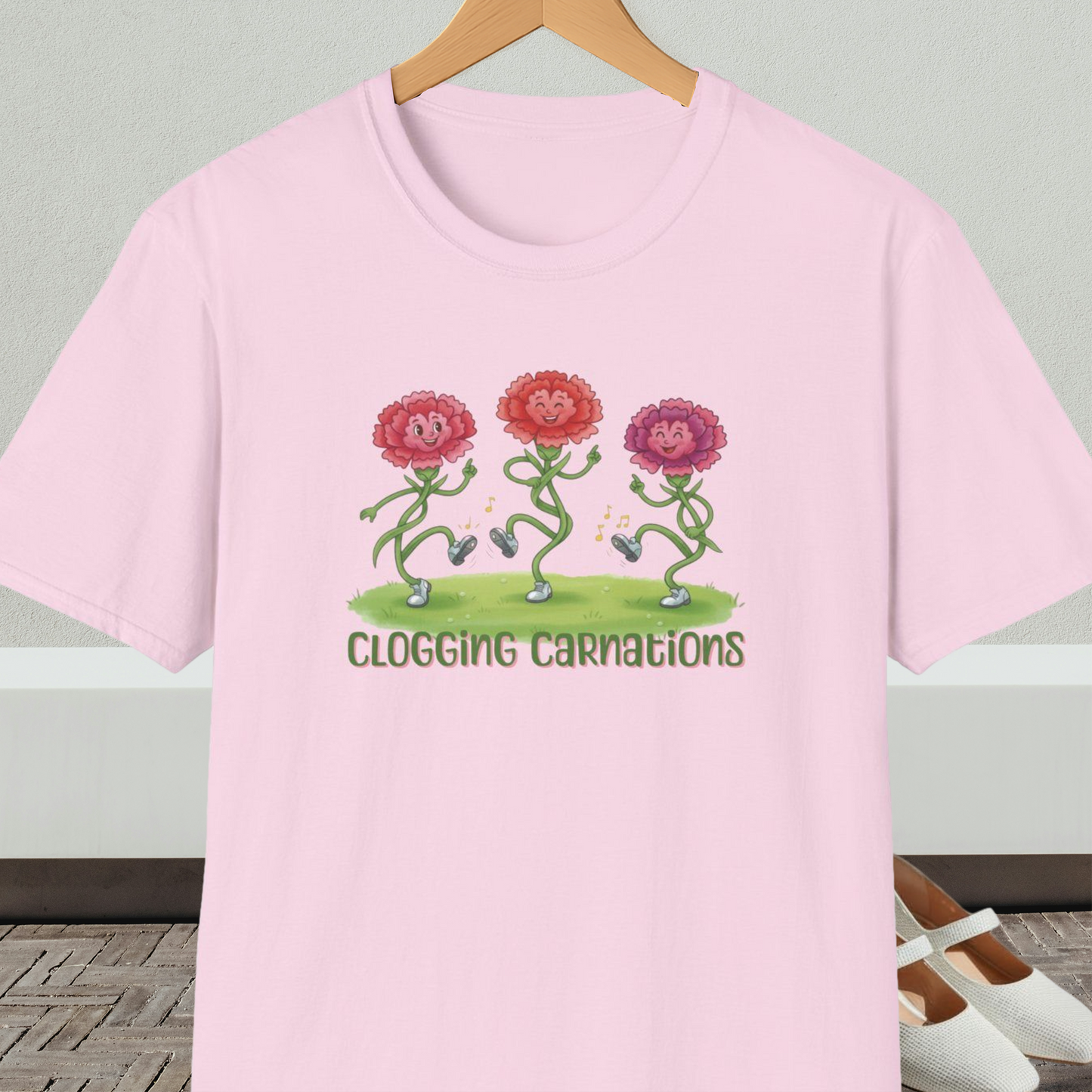 Light pink t-shirt with dancing flower design and text 'Clogging Carnations', on a hanger in front of a neutral background.