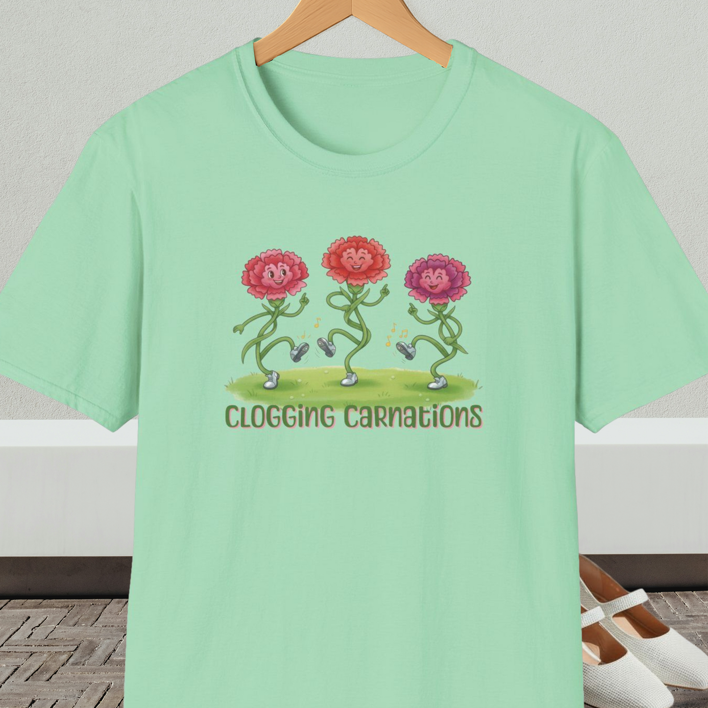 Light green t-shirt with dancing flower design and text 'Clogging Carnations', on a hanger in front of a neutral background.